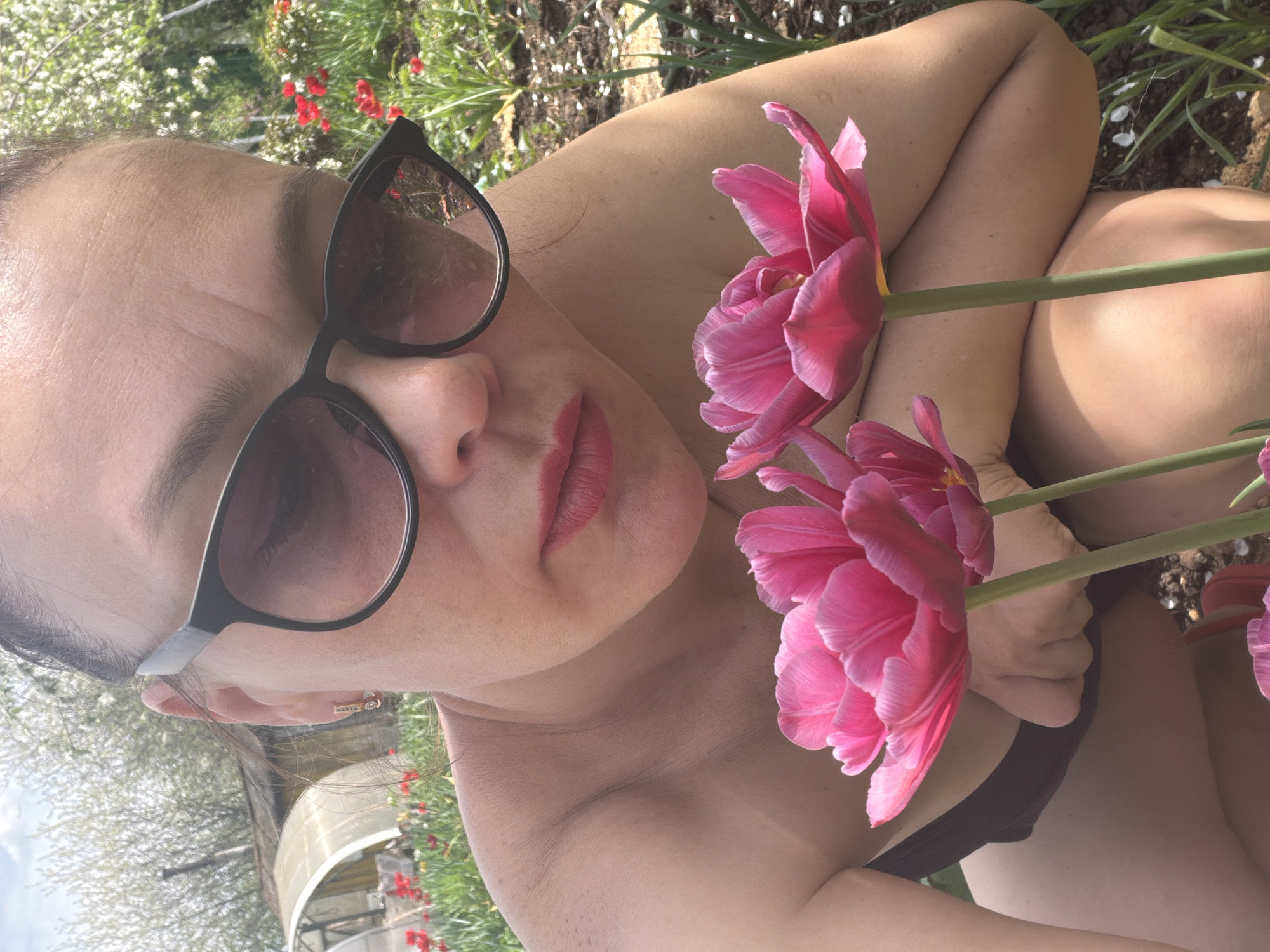 A woman wearing sunglasses and lipstick is lying on the ground next to pink tulips. Her arms are crossed. She is wearing a black bikini top. There is vegetation around her. A building can be seen in the background. The location is Chaikovsky, Russia.