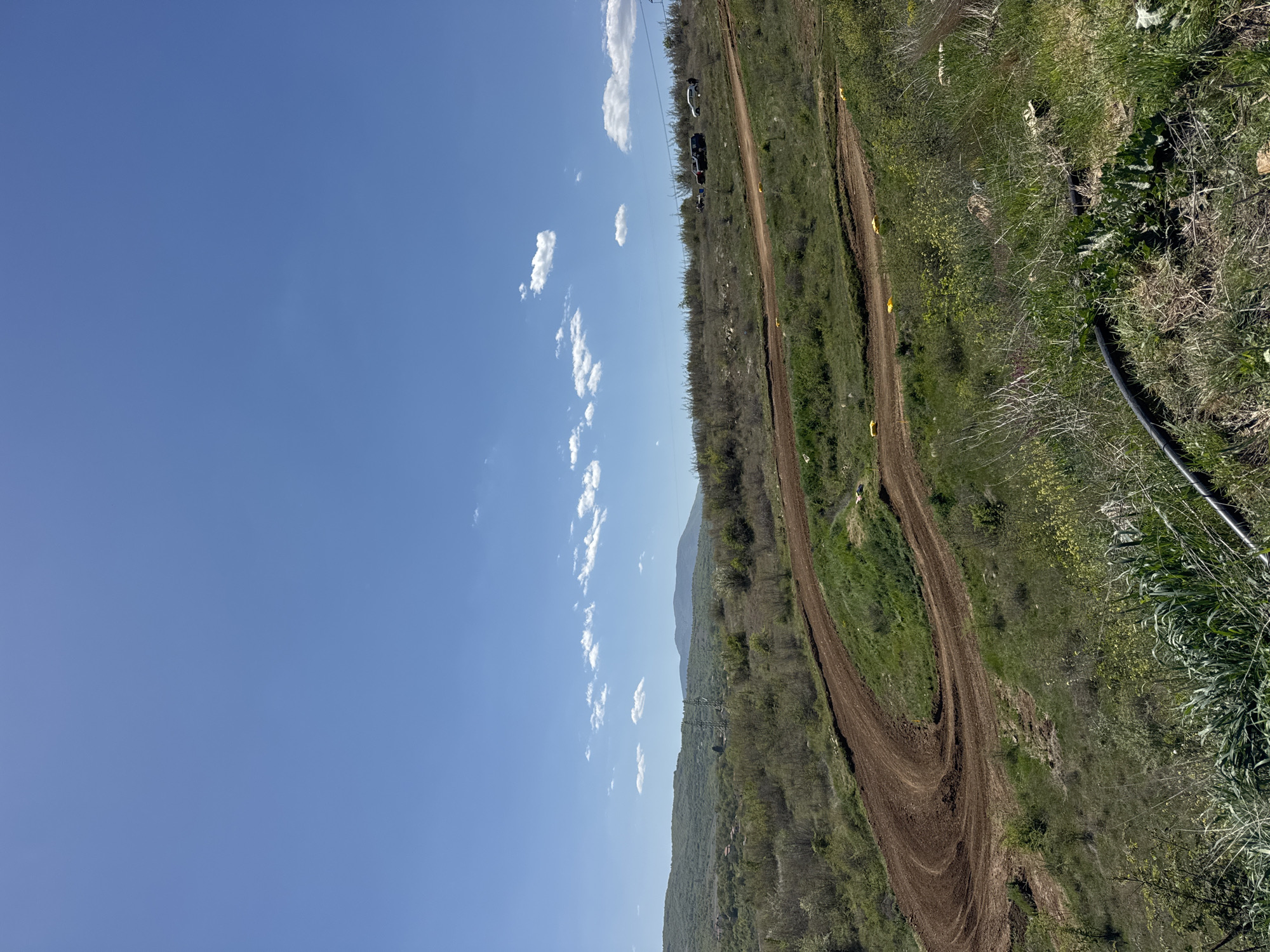 The image shows a dirt track in Selishte, Bulgaria, surrounded by green vegetation. The track curves through the landscape. The sky is blue with scattered clouds. There are power lines and vehicles in the background. The terrain appears hilly and mountainous. There is a black object near the bottom right corner of the image.