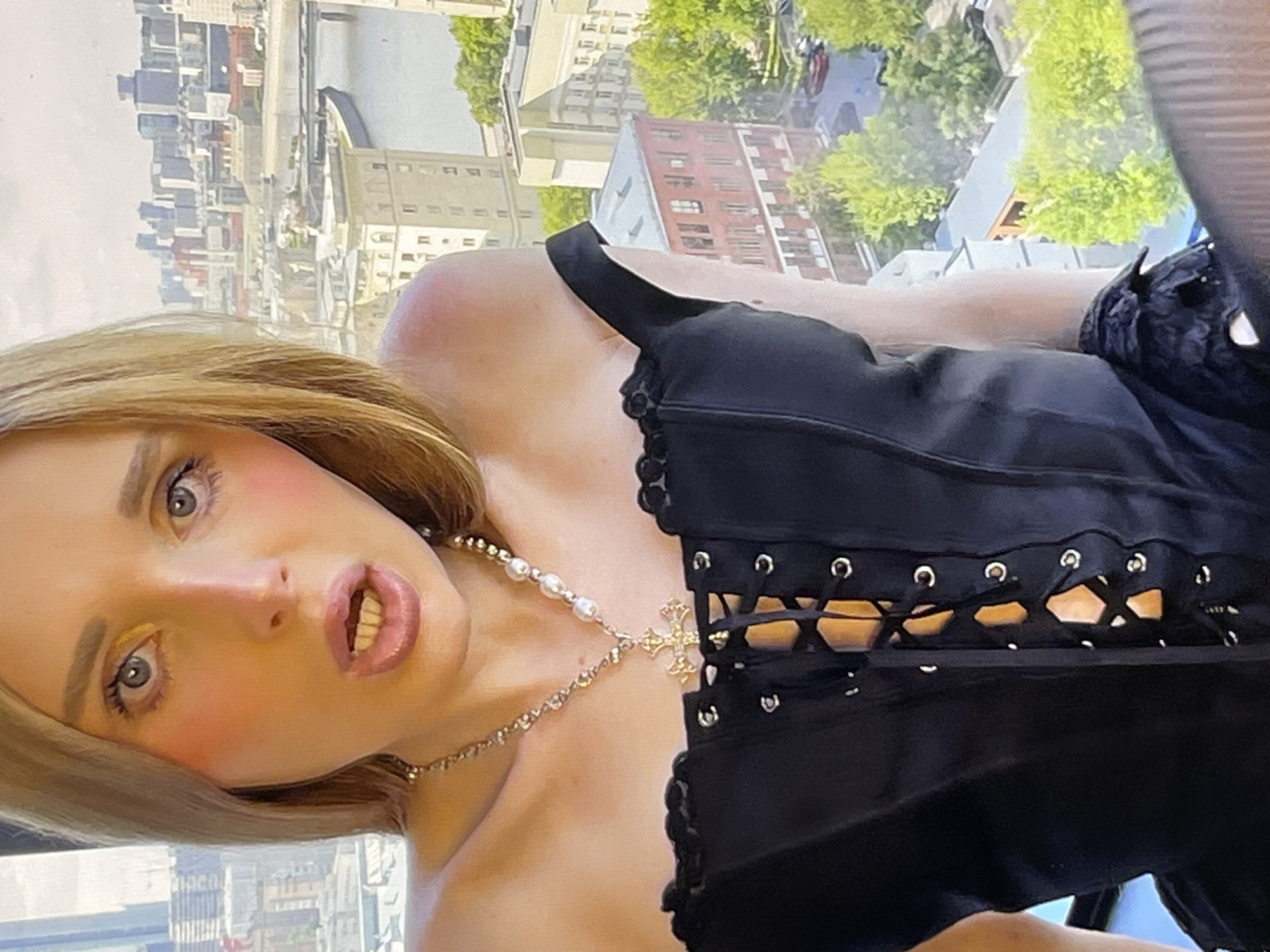 This media file shows a close-up, head-and-shoulders shot of a person with blonde hair and light blue eyes, wearing makeup that includes blush on the cheeks and eyeshadow. They are looking directly at the camera with their mouth slightly open, revealing their teeth. Around their neck, they are wearing a pearl necklace with a golden cross pendant. The person is wearing a black corset with lace-up detailing and what appears to be lace trim along the top.
The background suggests an urban setting with a view of buildings and trees, hinting at an outdoor or elevated location, possibly a city. The lighting suggests it is daytime. The image is oriented sideways. There is no visible text in the image.