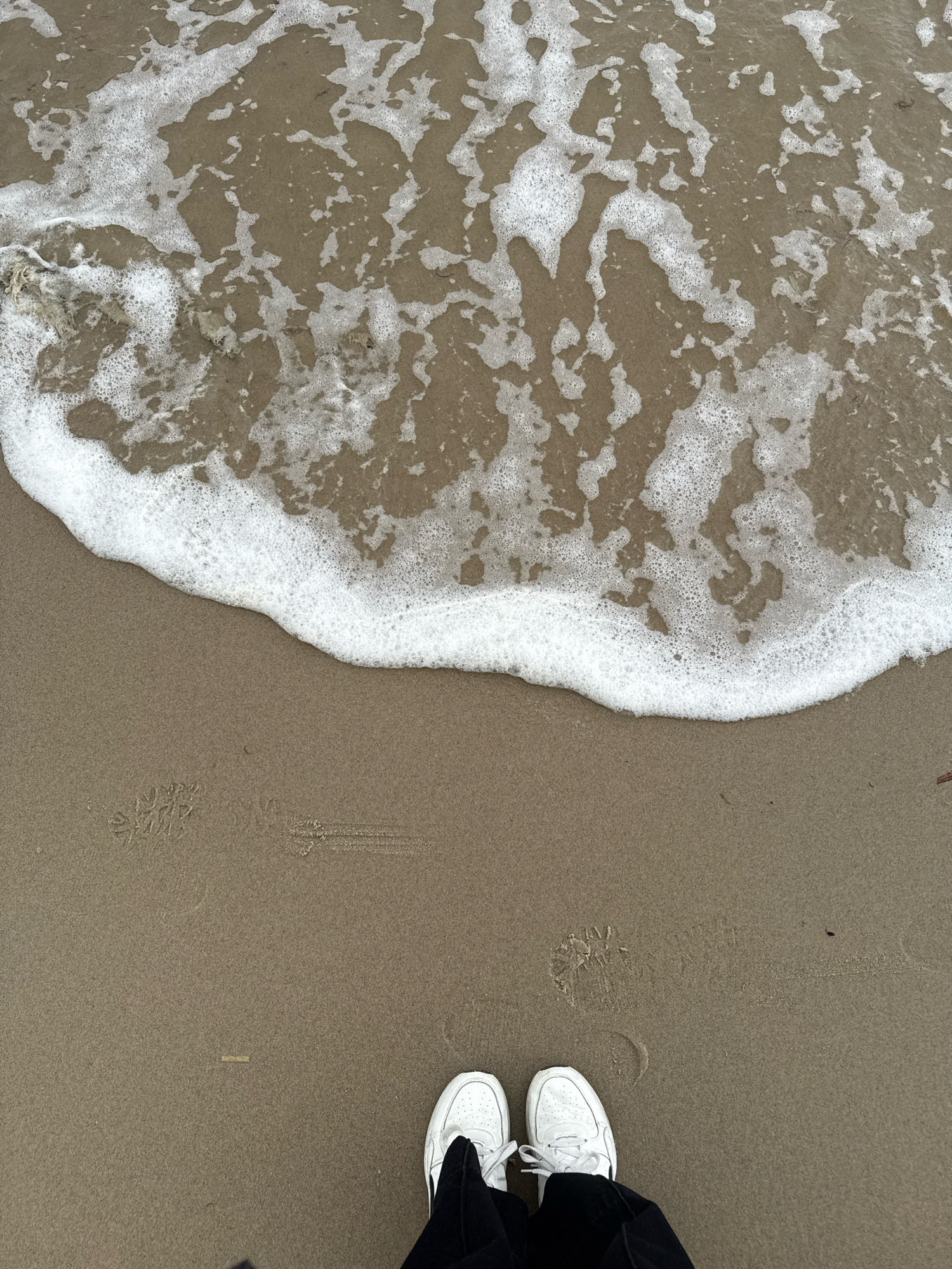 The image shows a top-down view of a sandy beach with gentle waves and foam washing ashore. In the foreground, a person's feet are visible, wearing white sneakers and black pants. There are footprints and bird tracks in the sand. The environment appears to be a coastal area. Based on the provided location information, this scene is set in Svetlogorsk, Russia.