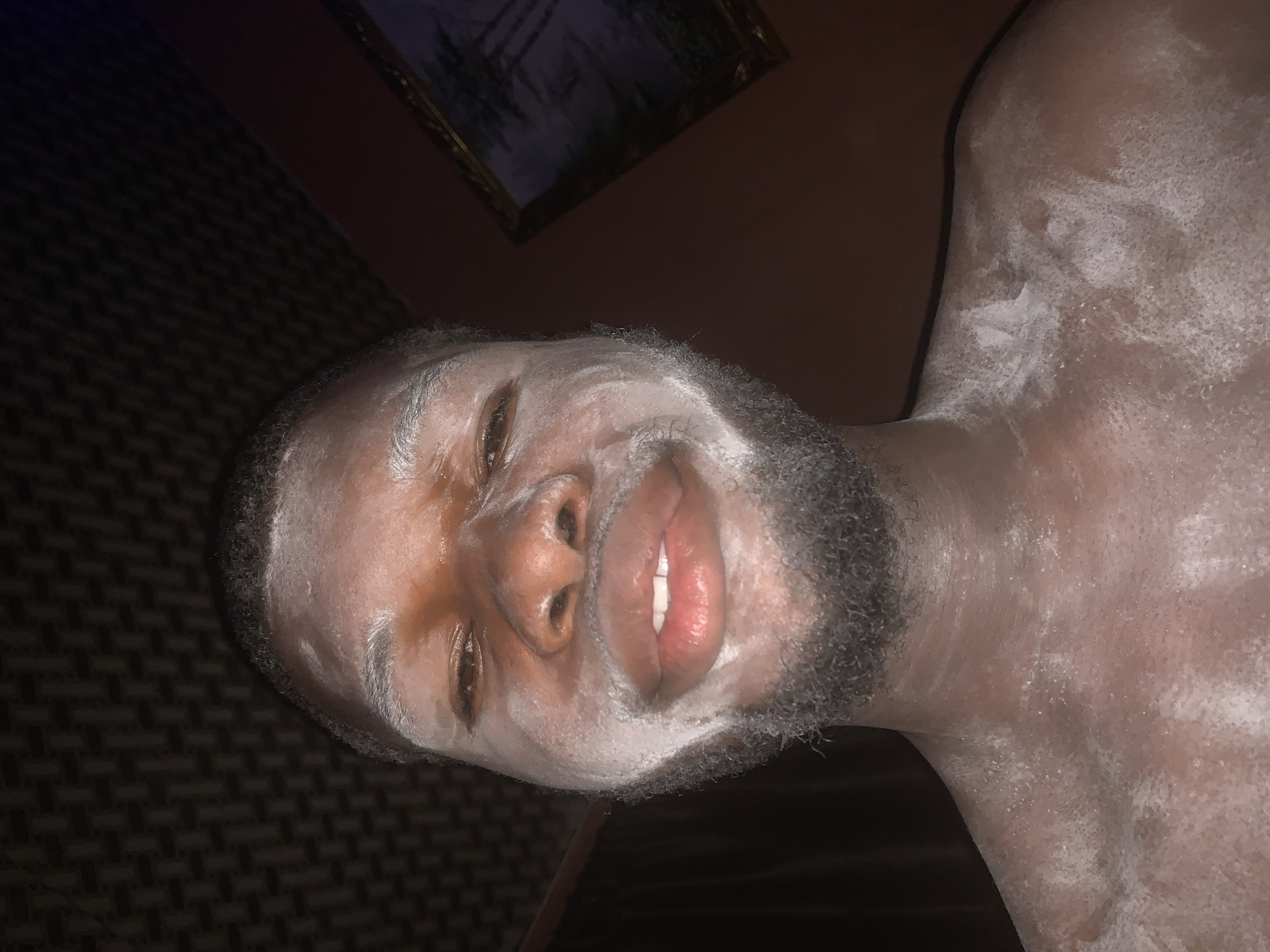 A close-up, rotated image depicts a Black man with a white, powdery substance applied to his face, neck, and upper chest. The substance appears particularly concentrated on his forehead, cheeks, jawline, and shoulders, partially covering his short, dark beard and hair on his head. His mouth is slightly open, revealing white teeth and pink lips, and his eyes are open, looking directly towards the viewer with a neutral to slightly smiling expression.
The man's skin tone is dark brown, and the white substance contrasts sharply against it. The lighting is dim, suggesting an indoor setting. In the background, to the left, there is a dark, textured surface, possibly a carpet or fabric wall hanging with a geometric pattern. To the right, the background is darker and less distinct, with a rectangular framed picture visible in the upper right corner. The details of the picture within the frame are obscured by darkness and blur.
The image does not contain any discernible text. The overall scene suggests a moment captured indoors, potentially during a personal grooming routine involving a face mask or body treatment, or perhaps as part of a costume or celebratory custom. Given the provided context, the location is Onitsha, Nigeria, though this is not visually evident in the image itself.