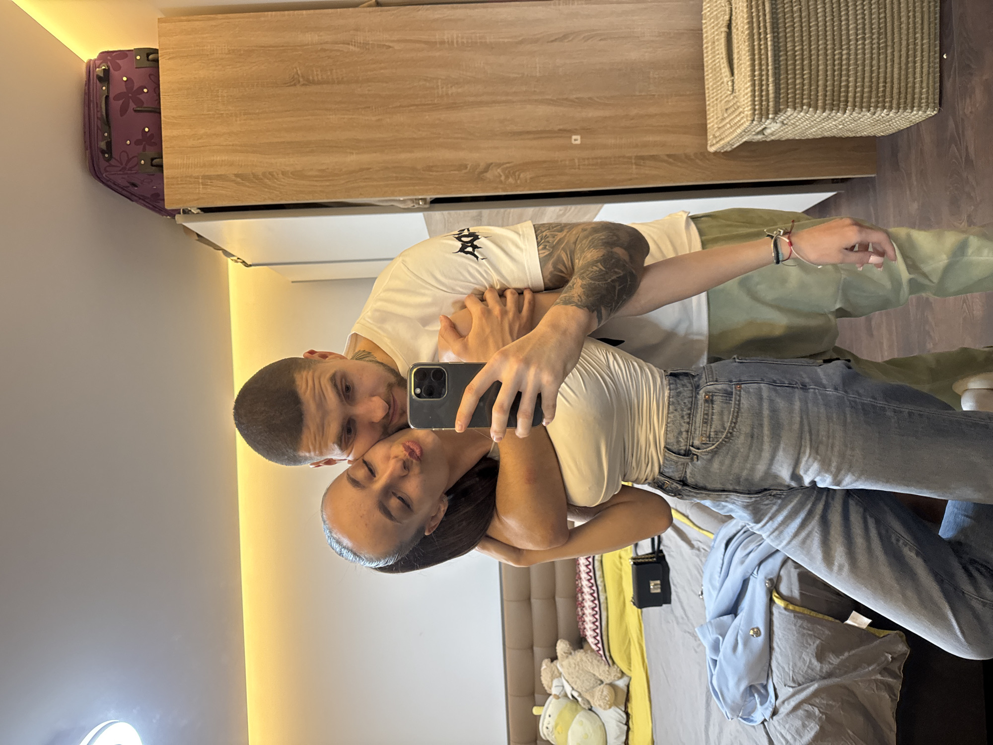 A mirror selfie captures a male and a female embracing within an indoor domestic setting. No visual identifiers are present to confirm the location as Sofia, Bulgaria. The female, positioned in front and slightly left, has dark hair in a ponytail, wears a white sleeveless top and light blue jeans. She holds a dark-colored smartphone in her left hand, taking the photograph. Her eyes are closed, and her lips are pursed. Her right arm is around the male's neck. The male, positioned behind her, has short dark hair and visible tattoos on his right arm. He wears a white short-sleeved t-shirt with a black graphic and olive green pants. His right arm is around her waist, and his left arm is around her upper arm. The background, reflected in the mirror, includes a bed with a brown padded headboard, grey bedding, a yellow blanket, a patterned pillow, and two stuffed animals. A black rectangular purse and a light blue garment with two silver buttons are also on the bed. A light wood-grain wardrobe is visible to the right, topped by a purple floral-patterned wheeled suitcase. A woven beige basket with a lid sits on a dark wood floor in the lower right. The room features light-colored walls, a white ceiling with a recessed light fixture, and a strip of warm ambient lighting along the ceiling's perimeter.