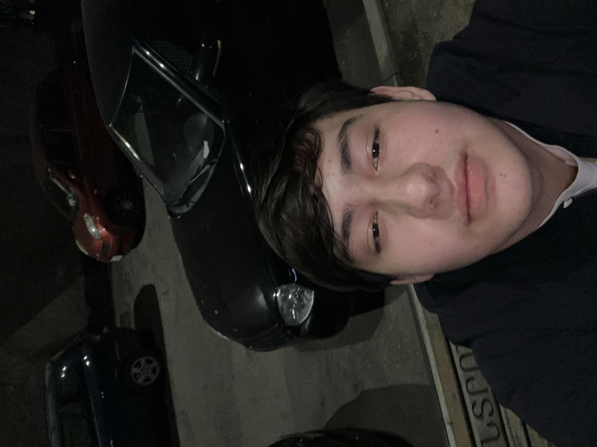 A person stands outdoors at night next to a dark-colored car with its headlights off. Several other cars, one red and one dark blue, are parked nearby. The person is fair-skinned and wearing a dark top. The location includes sidewalk markings.