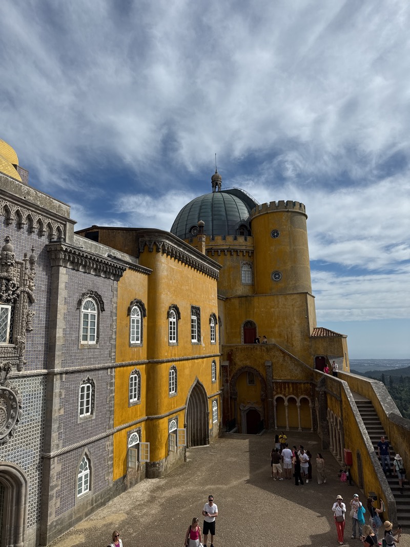 The image captures a high-angle view of Pena Palace in Sintra, Portugal, under a sky with partly cloudy formations. The architectural structures display a mix of colors, including yellow, gray, and touches of gold. The yellow section of the palace features windows with white frames, and an arched entrance. The gray section is adorned with patterned tiles and decorative window frames. A rounded tower rises behind the yellow section, capped with a green dome and a small spire.
A courtyard is visible in the center of the frame, where a group of people are gathered. Individuals are seen walking and standing, some of them in casual clothing. A staircase runs along the side of the building, with more people ascending or descending it. The courtyard itself is paved. A glimpse of the surrounding landscape is visible in the distance.