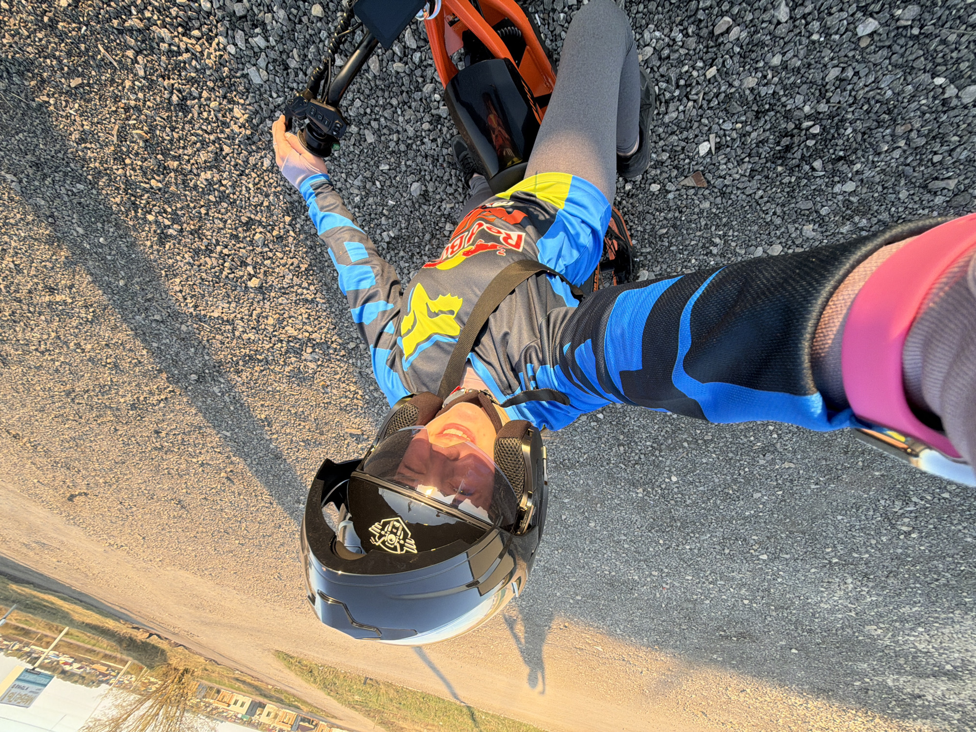 A person wearing a black motorcycle helmet, a blue and grey long-sleeved shirt with "Red Bull" and "FOX" logos, and grey leggings is lying on their back on a gravel surface. The person is positioned on or near an orange and black electric scooter or small motorbike. The individual appears to be holding a smartphone or camera to take a selfie. In the background, there is a glimpse of a cityscape, possibly indicating the location of Азьмушкино, Russia. The scene suggests an outdoor setting, possibly a roadside or recreational area.