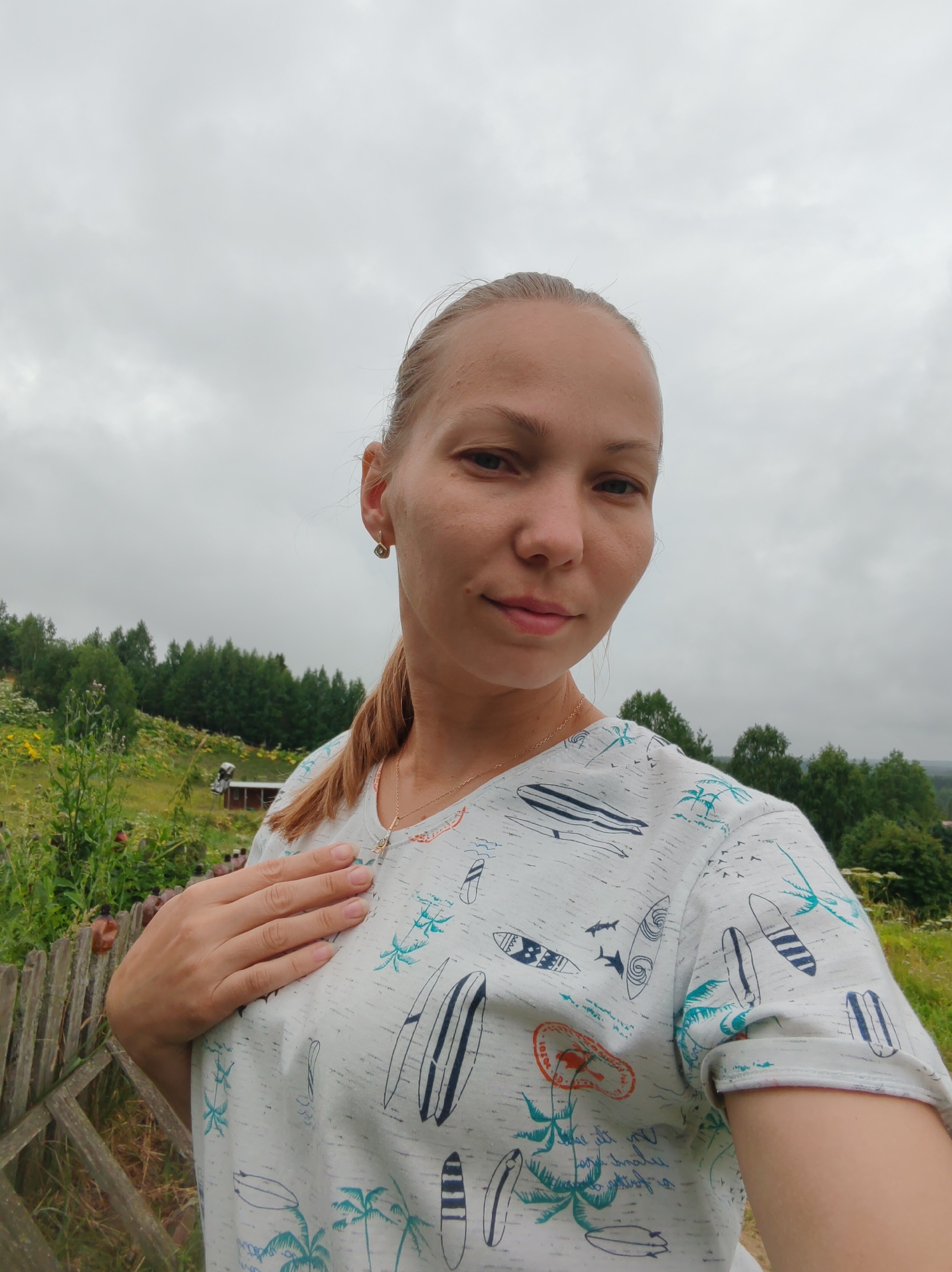 The image is a selfie taken outdoors. A woman with blonde hair pulled back in a ponytail is the primary subject. She is wearing a light-colored t-shirt with a tropical print featuring surfboards and palm trees. She has one hand placed on her chest. The background consists of a wooded hillside under a cloudy sky. There is a wooden fence in the foreground and some vegetation, including wildflowers, on the hillside. The location is identified as Chukhlem, Russia.