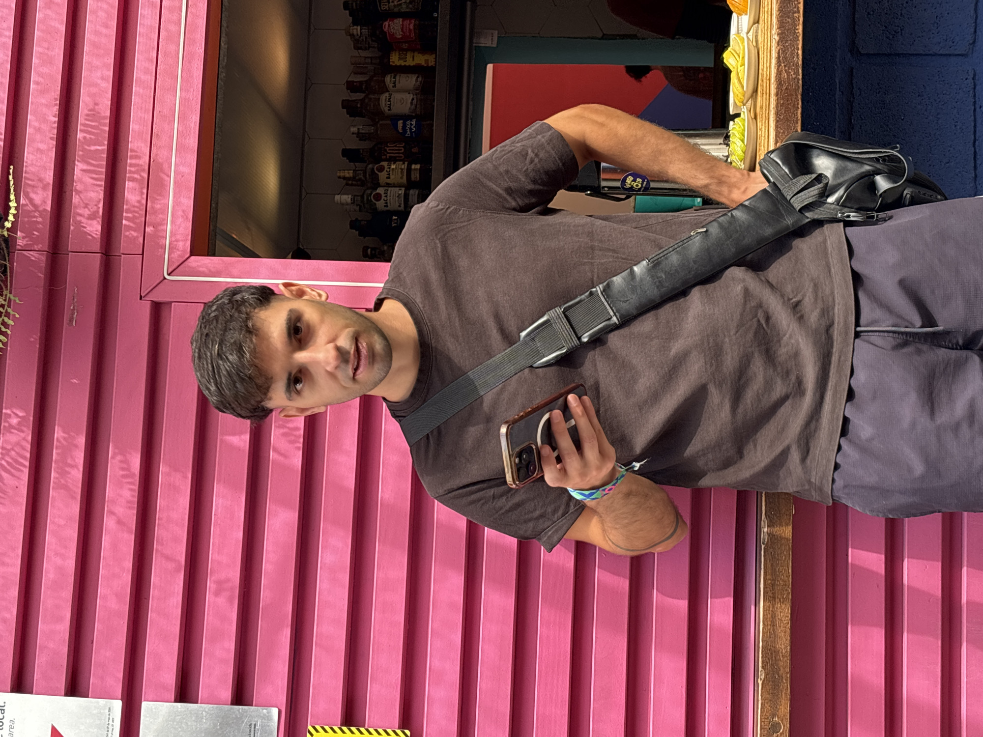 A man stands in front of a pink corrugated wall, holding a smartphone in his left hand. He wears a brown t-shirt, dark blue pants, and a black bag across his torso. The man has dark hair and is looking at the camera. Behind him is a partially visible bar area with bottles and other items on display. The bar appears to be in a location in the city of São Paulo, Brazil.