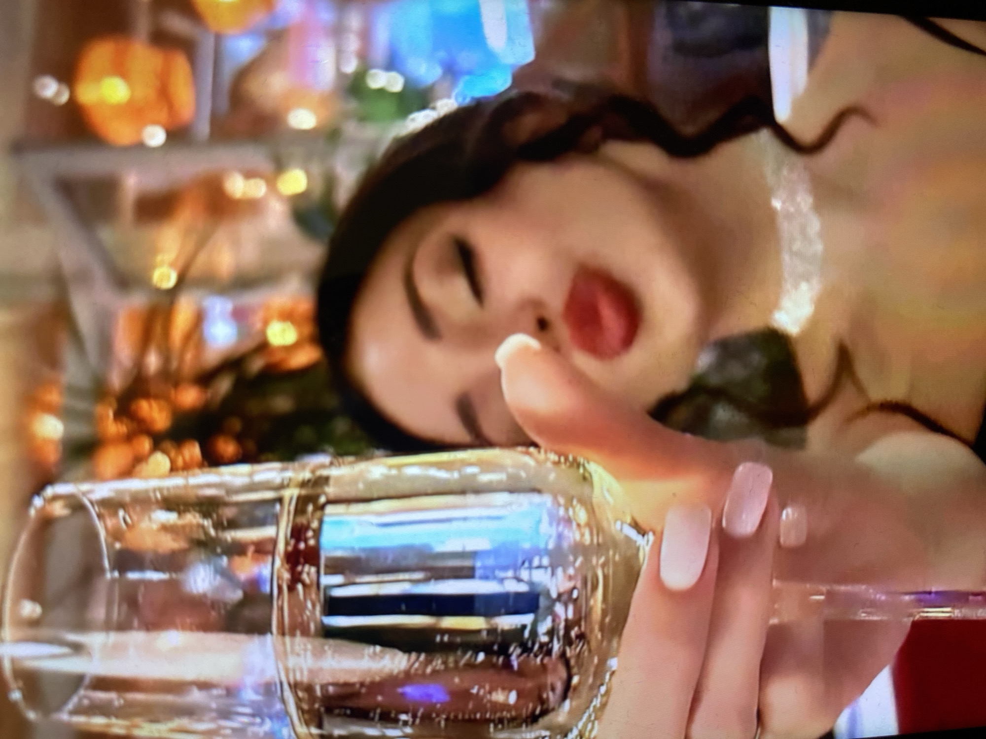 A woman with dark curly hair and bright red lipstick is captured in a close-up shot, holding a champagne flute filled with a bubbly beverage. She is leaning forward, her lips pursed as if to kiss the camera, and her eyes are closed. She wears a delicate white choker necklace around her neck. The background is softly blurred, revealing warm, orange-toned lights, suggesting an indoor evening setting, possibly a restaurant or bar. The image's quality is grainy, typical of a photograph taken on a screen or from a distance. There is no information to indicate the location is Kamianske, Ukraine.