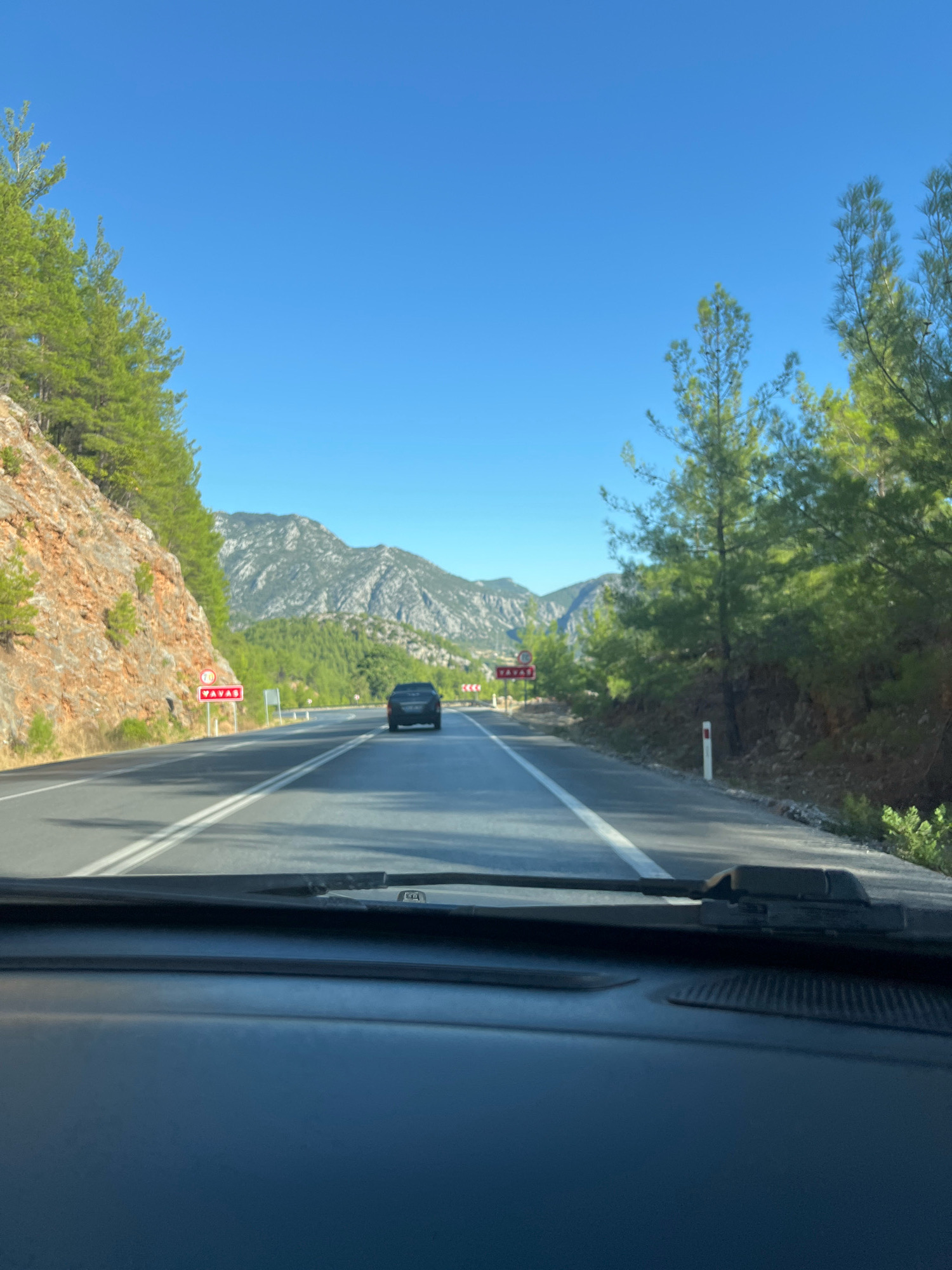 The image shows a view from inside a vehicle looking forward on a paved road with a clear blue sky overhead. The road has double white lines in the center and extends into the distance towards mountains. On the left side of the road, there is a rocky cliff face with pine trees growing on it. On the right side of the road, there are more pine trees and vegetation. A black car is driving ahead on the road, and there are several road signs visible, including one with a speed limit of 70. The location is identified as the Akseki-Konya highway in Akseki, Turkey, a region known for its mountainous terrain and pine forests. The scene depicts a typical travel view on a sunny day in this area.