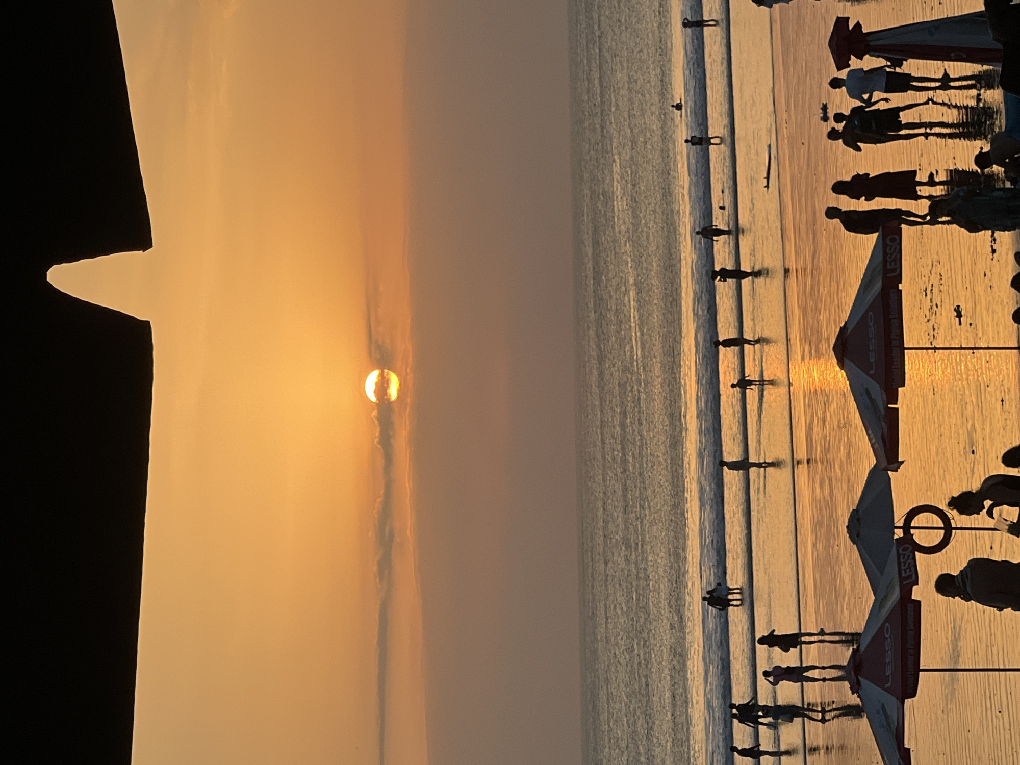 The image depicts a sunset scene at Kuta Beach in Kuta, Indonesia. The sun, a bright orb, is partially obscured by clouds, casting a warm orange glow across the sky and the ocean. Silhouettes of people are visible on the wet sand and in the shallow water, some walking, some standing, and some interacting. Several beach umbrellas, branded with "LESSO", are set up in the foreground. The calm ocean waves gently lap the shore, reflecting the sunset's colors. The overall atmosphere is one of leisure and tranquility, typical of a beach at dusk.