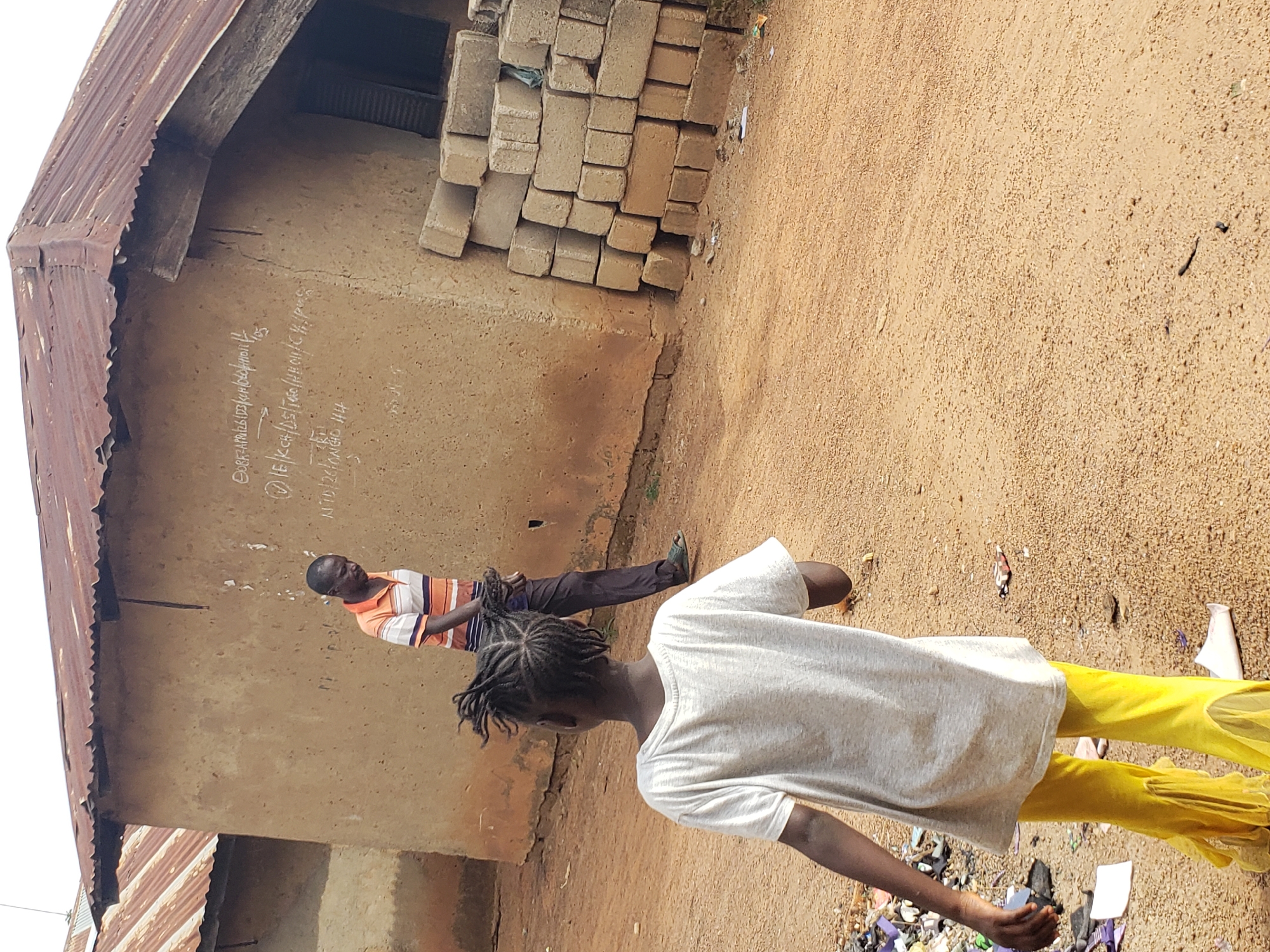 In Kachia, Nigeria, a dirt path leads towards a compound with buildings constructed of mud walls and corrugated metal roofs. In the foreground, a young girl with braided hair, wearing a grey t-shirt and bright yellow pants, walks away from the viewer. Behind her, a man in an orange and striped t-shirt and dark pants is visible, seemingly carrying or interacting with a bundle of dark, coiled material. To the right of the buildings, a stack of concrete blocks is piled against the wall. The ground is sandy and dry, with scattered debris. White writing is visible on the mud wall of the building.