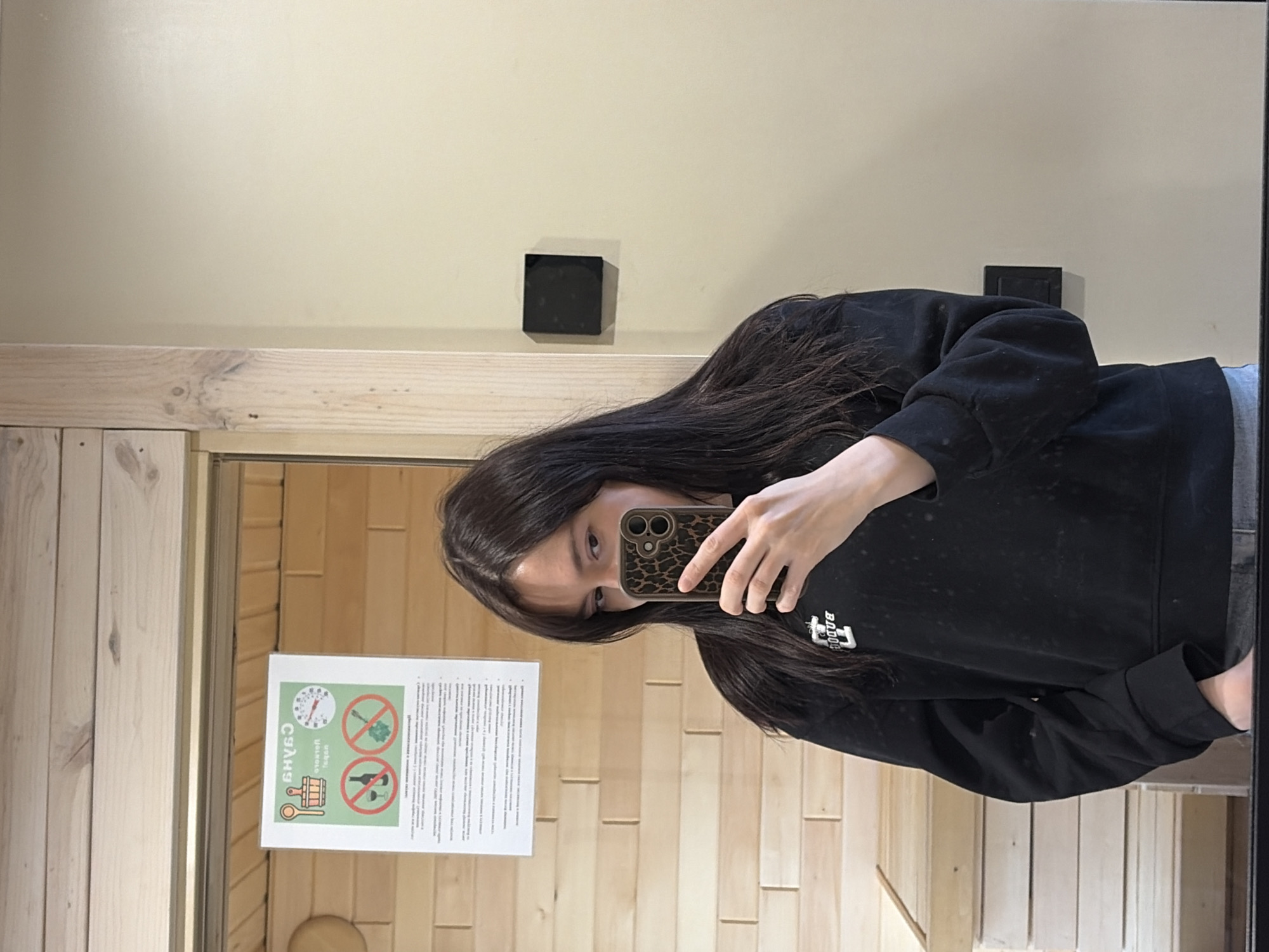 A person is seen taking a mirror selfie indoors. The subject is a female with long, dark hair, wearing a black hooded sweatshirt with a white logo (partially visible, possibly "BOJO"). She holds a smartphone with a distinctive leopard print case, obscuring her mouth and chin as she takes the photo, with her eyes looking towards the camera.
The setting appears to be a room, possibly within a spa or sauna facility, given the prominent light-colored wooden paneling on the wall, which has a distinct block pattern. Above the wooden paneling, the wall is painted a light, neutral beige color. A small, black rectangular object, possibly an electrical outlet or light switch, is mounted on the beige wall.
Reflected in the mirror, to the left of the person, is a white rectangular sign attached to the wooden wall. The sign features Cyrillic text at the top, including "САУНА" (SAUNA), confirming the location context as a sauna. Below the text are several pictograms: a thermometer, a bucket with a ladle, a crossed-out bath broom (venik), and a crossed-out bottle, likely indicating rules or recommendations for sauna use. The overall ambiance suggests an indoor, clean environment. The location is Мишкино, Russia, consistent with the Cyrillic text on the sign.