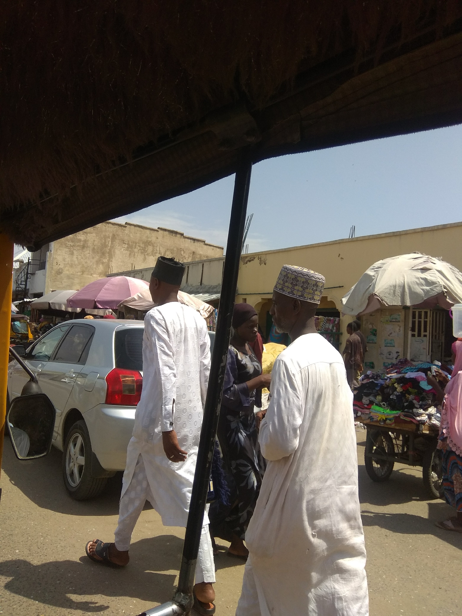 In Jalingo, Nigeria, a group of individuals are captured in a bustling street scene. Two men in traditional white attire, one wearing a black cap and the other a patterned cap, walk side-by-side. Behind them, a woman in a dark patterned dress is visible. In the background, a silver sedan and a rickshaw are parked on the dusty street, suggesting a busy marketplace or thoroughfare. Buildings with light-colored walls and a market stall adorned with clothing are also part of the urban landscape. The scene is illuminated by bright sunlight, casting distinct shadows on the ground.