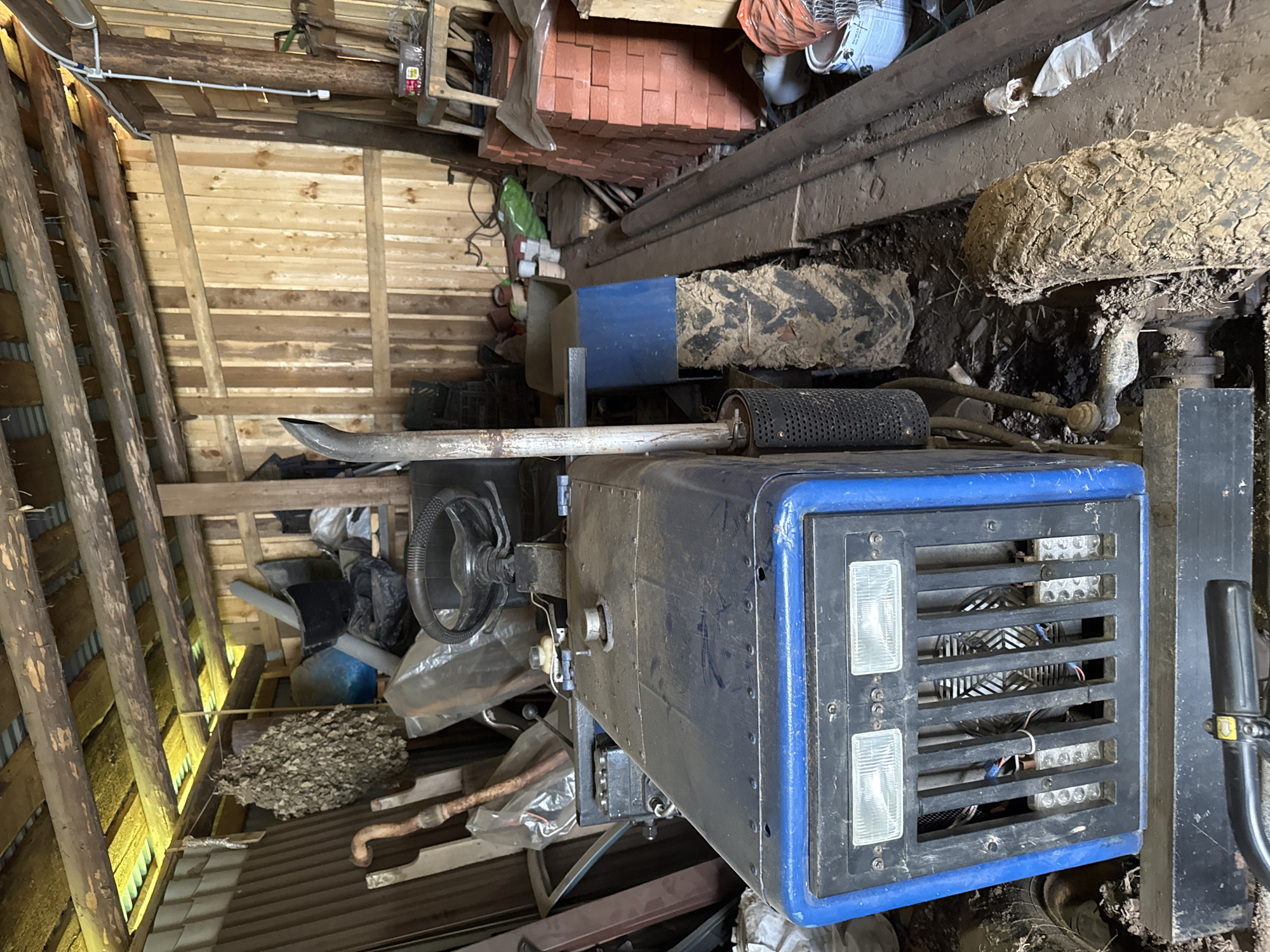 The image depicts the interior of a rustic wooden shed in Бородулино, Russia. The shed is filled with various items, suggesting storage and possibly agricultural use. Dominating the foreground is a blue and black tractor, its front grill featuring two rectangular headlights and a fan visible behind a metal grille. The tractor is dirty, with mud caked onto its tires. To the left of the tractor, a collection of dried branches, possibly a bathhouse broom (venik), hangs from the ceiling. Various tools and implements, including a curved metal object resembling a handle, are leaning against the walls. The shed's construction is visible, with rough-hewn wooden planks forming the walls and ceiling, supported by wooden beams. The floor is cluttered with debris, including dirt, straw, and discarded materials. In the background, stacks of red bricks and buckets are visible, along with other indistinct objects. The overall impression is one of a functional, albeit somewhat disorganized, storage space in a rural setting. There are no people present in the image.