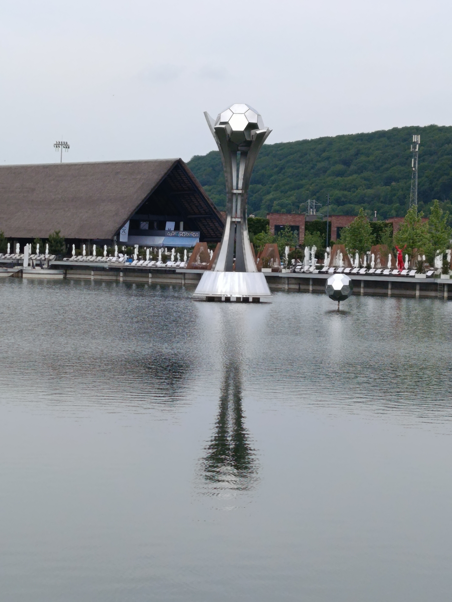 This image presents an outdoor scene featuring a large, still body of water, likely an artificial lake or pond, at a recreational area in Vynnyky, Ukraine. The central and most prominent subject is a tall, modern, silver-colored sculpture resembling a large trophy, with a faceted spherical top. This sculpture is positioned within the water and its reflection is clearly visible on the surface.
To the right of the main sculpture, a smaller, similarly faceted silver sphere floats on the water. On the left bank, a large building with a distinctive dark, steeply pitched roof (possibly thatched or shingled) is visible. In front of this building, along the water's edge, a deck is lined with numerous white sun loungers or chairs. A banner with faint, illegible blue text hangs on the building's facade.
On the right bank, another section of deck features more white loungers, some covered, and several brown, open-lattice structures, possibly pergolas. A single figure, clad in a striking bright red outfit, stands on this bank with arms raised, providing a strong point of color against the otherwise subdued palette.
The background consists of dense, green forested hills under an overcast, light grey sky, suggesting diffused daylight. A cellular tower stands on the right side of the image, emerging from the hillside. The water's surface shows subtle ripples and reflects the surrounding elements. The scene appears to depict a modern leisure facility or resort.