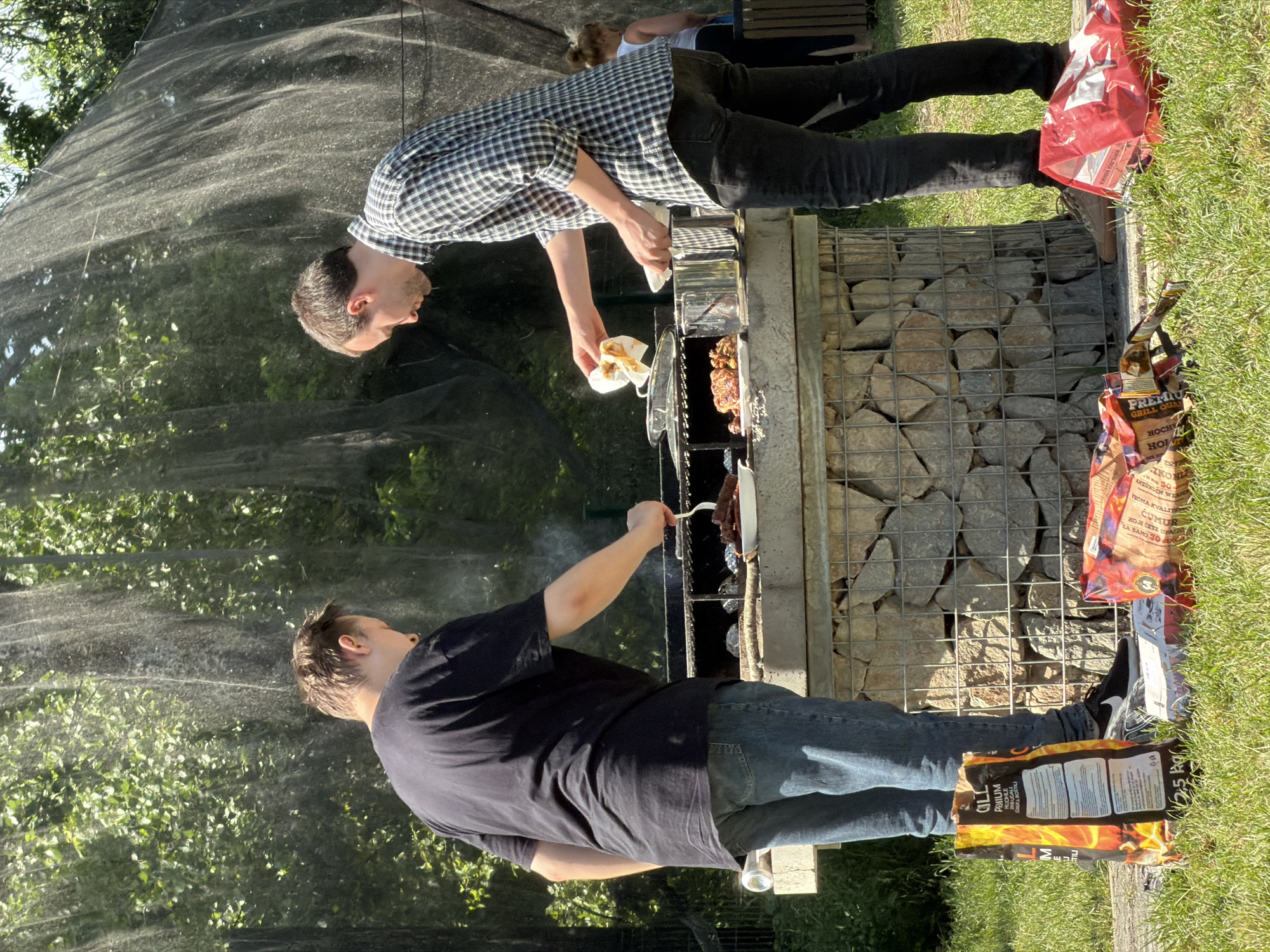 Two men are barbecuing outdoors, seemingly in the city of Cukarica Urban Municipality, Serbia, due to the writing on the charcoal bag. One man, wearing a black shirt and blue jeans, is using a fork to turn meat on the grill. The other man, in a checkered shirt and black jeans, is holding a plate and wiping it with a white cloth. The grill is made of stone and metal mesh, and bags of charcoal are on the ground nearby. The background includes a green, leafy area and a dark net-like structure. The scene is set in daylight, with the sun casting shadows on the environment.