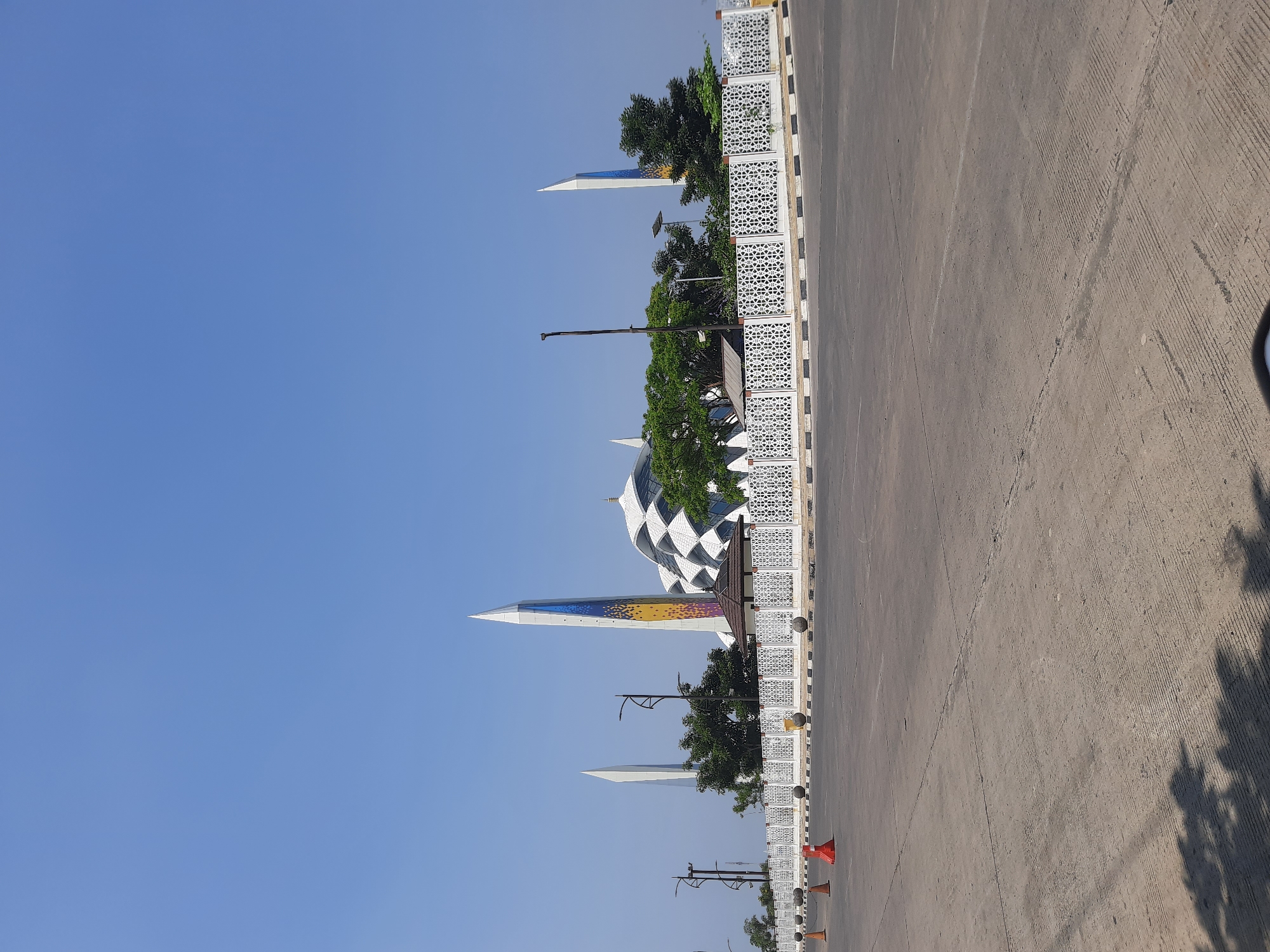 This image depicts the exterior of a modern mosque in Bandung City, Indonesia, under a clear blue sky. The mosque features a distinctive dome with a geometric pattern, two tall, slender minarets, one of which is decorated with colorful geometric designs, and another smaller minaret. A white decorative fence with an intricate geometric pattern runs along the front of the mosque, separating it from a paved area in the foreground. Several traffic cones and bollards are visible on the paved surface, suggesting a public space or parking area. Trees are planted behind the fence, and the overall scene is bathed in bright sunlight, casting shadows on the pavement.