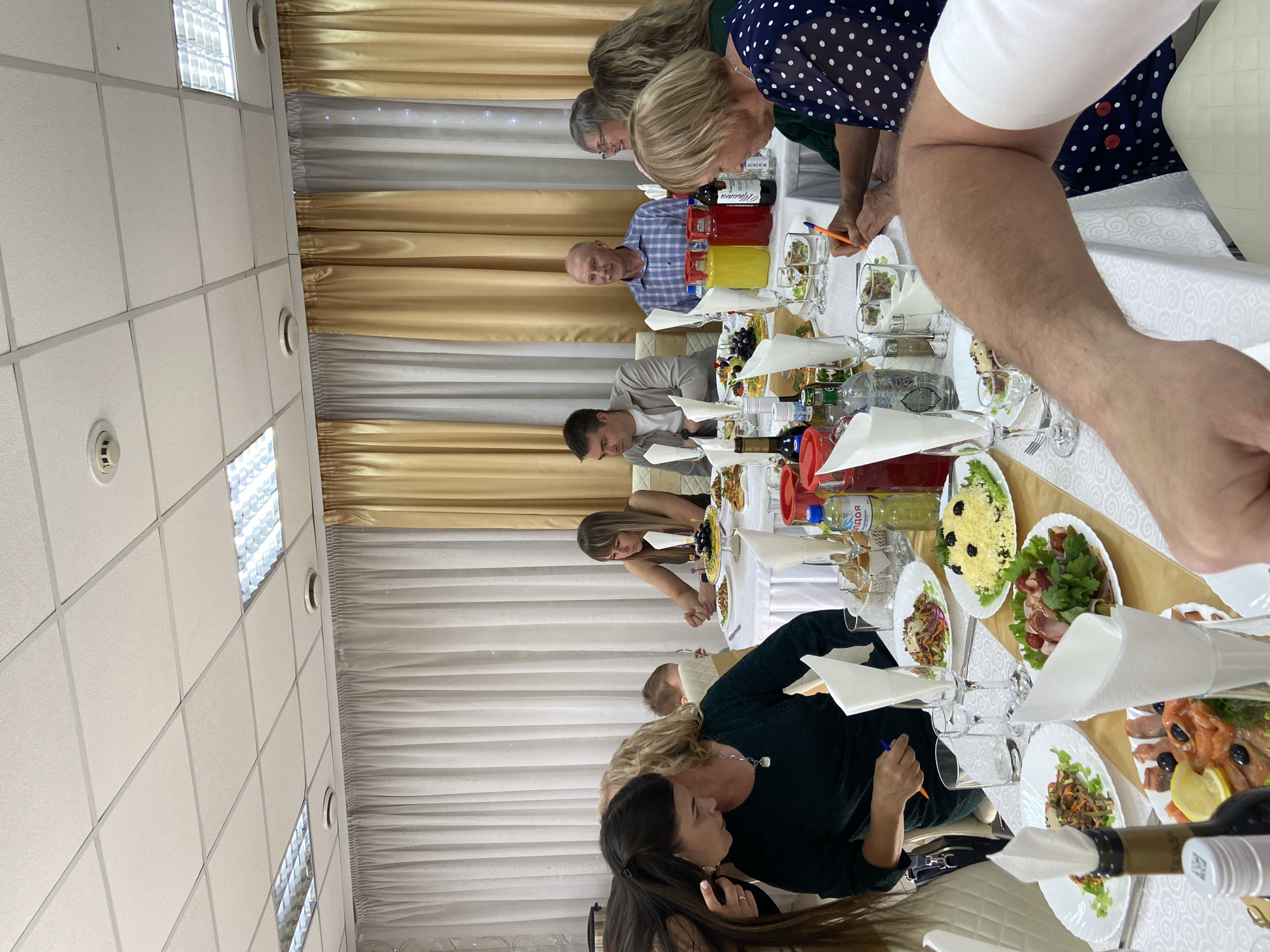 A group of people are seated at a long, white-clothed table laden with food and drinks. Curtains, half white and half gold, hang in the background. Plates of salads, seafood, and other dishes are visible, along with bottles of wine and juice. Some individuals are actively engaged in conversation or eating, while others appear to be observing. The setting suggests a formal gathering or celebration.