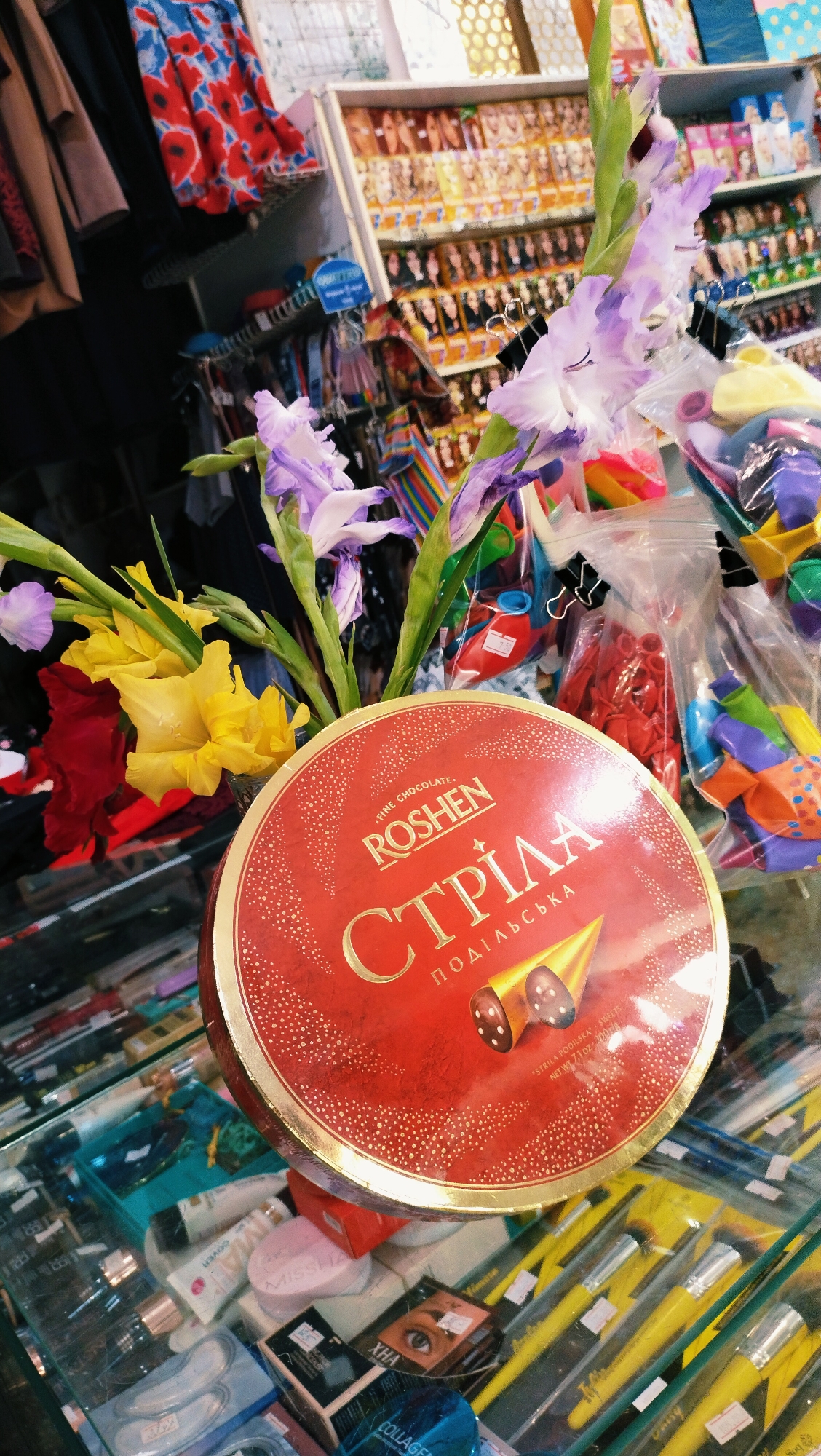 A high-angle medium shot shows a round, red chocolate tin with gold lettering placed on a glass display case filled with various items. The tin is adorned with the brand name "ROSHE N" and Cyrillic text. A bouquet of gladiolus flowers, with purple and yellow blooms, is arranged in front of the tin, partially obscuring it. The background reveals shelves stocked with colorful merchandise, including clothing and bags. Below the tin, the display case exhibits makeup and cosmetic products, with makeup brushes visible in the lower right corner. The overall setting appears to be a retail environment. Based on the specific product labeling ("STRILA PODILSKA"), this image is likely from a store in Horishni Plavni, Ukraine, known for its confectioneries.
