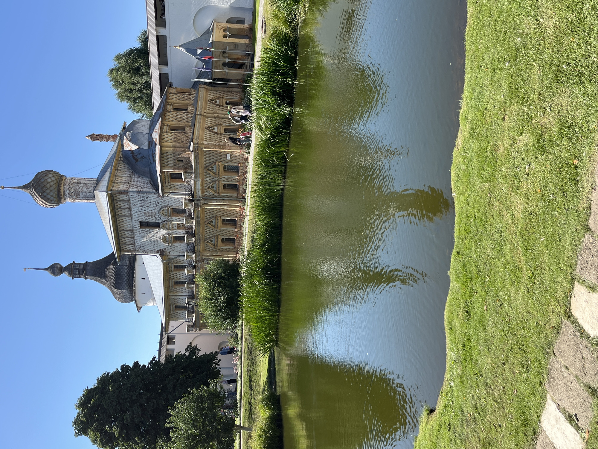 This is a bright, eye-level shot of a pond with a reflection of a building with multiple domes and spires, and a tall clock tower. The building is decorated with intricate patterns. In the foreground, there is a grassy bank with reeds growing near the edge of the pond. Behind the pond, there is a stone pathway and manicured grass. Several people are visible walking along the pathway near the building. The sky is clear blue and there are trees and more white walled buildings in the background. The scene is located in Novo-Nikolskoye, Russia.