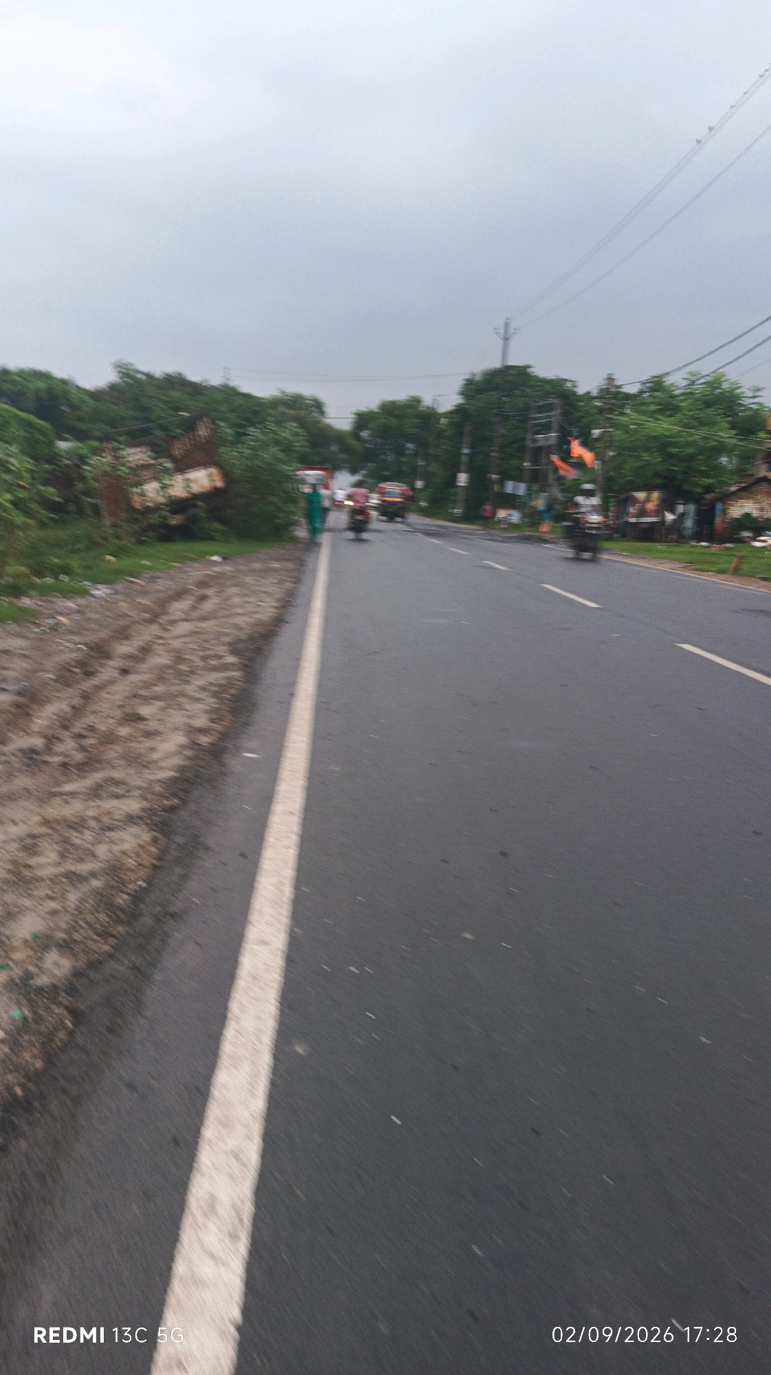A cloudy day is captured on a two-lane asphalt road. The road is bordered on the left by a sandy embankment covered in vegetation and on the right by trees and buildings. There are a few vehicles on the road, including a motorcycle, a car, and what appears to be an auto-rickshaw. One person in a green outfit is walking on the left side of the road, away from the camera. A discarded truck is visible on the left side of the frame, partially obscured by foliage. Orange flags are mounted on poles along the right side of the road. A date and timestamp, "02/09/2026 17:28," and "REDMI 13C 5G" are present in the bottom right and left corners of the image, respectively.