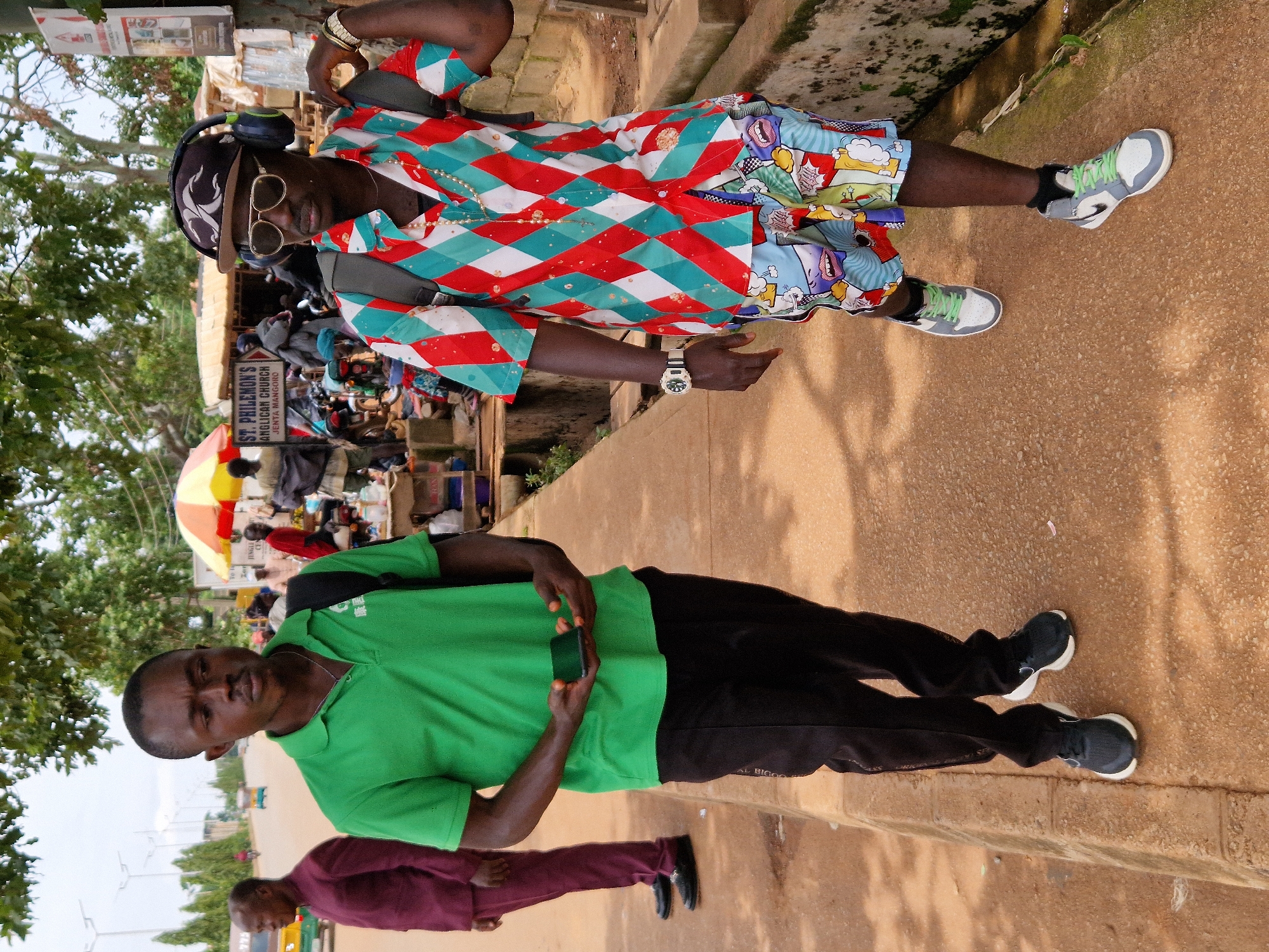 In an outdoor setting in Unknown, Unknown, two Black men walk on a paved sidewalk next to a dirt road. The man on the left wears a green polo shirt and black pants, holding a smartphone in his hands. The man on the right sports a colorful, diamond-patterned short-sleeved shirt, patterned shorts, sunglasses, headphones, and a watch, carrying a backpack. Several other individuals and a kiosk with a colorful umbrella are visible in the background, along with parked motorcycles and vehicles. The environment appears to be a bustling street scene.