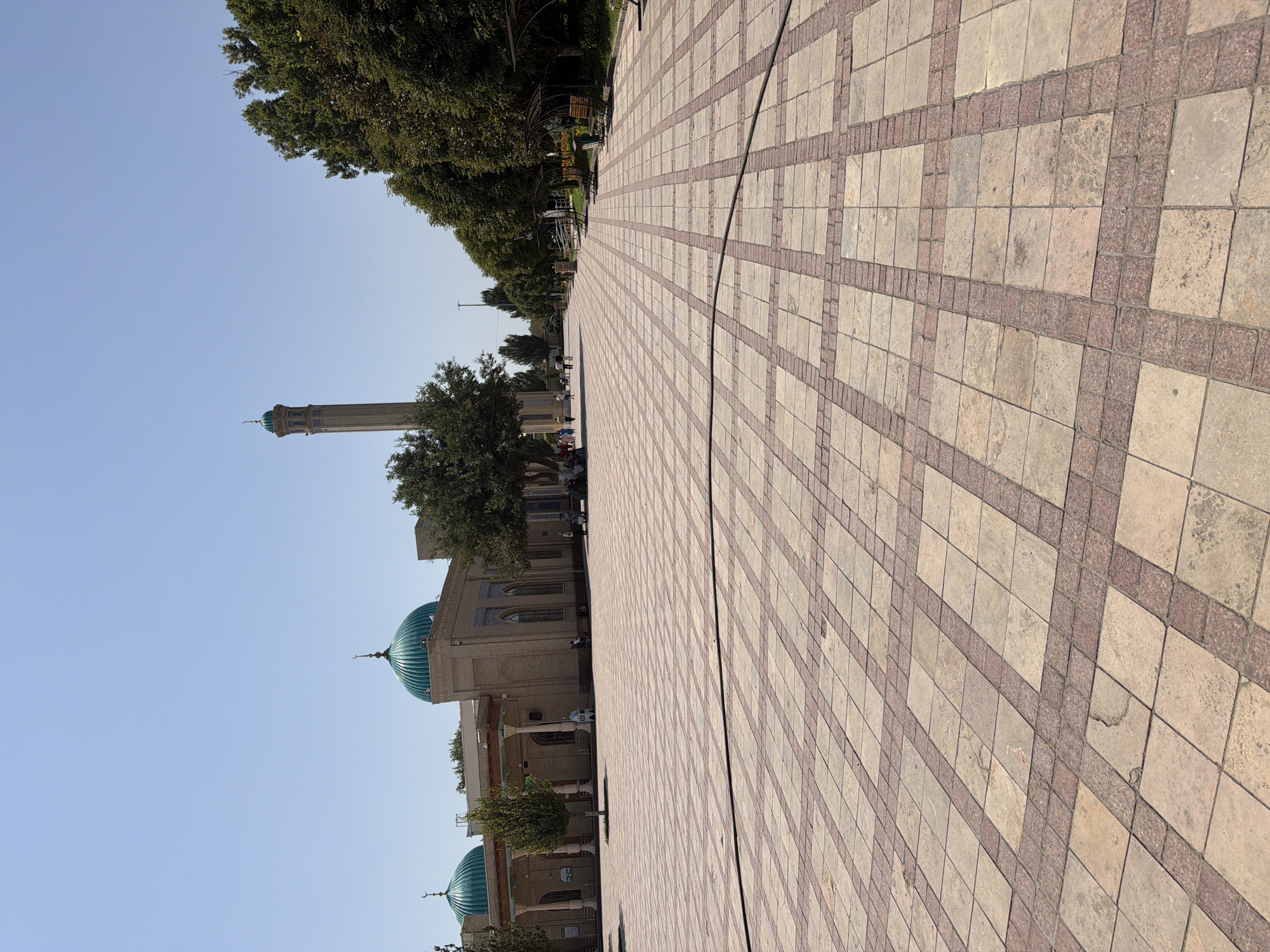 The image depicts a wide, paved courtyard leading towards a mosque in Zangiata, Uzbekistan. The courtyard is tiled with large, light-colored square tiles arranged in a diamond pattern, bordered by smaller, darker reddish-brown tiles. A black hose or cable runs diagonally across the foreground.

To the left of the courtyard, a tan-colored mosque with two prominent turquoise domes and a tall, slender minaret is visible. The mosque has arched windows and decorative blue tilework on the minaret. Several trees, including a large one in front of the mosque, provide shade.

On the right side of the courtyard, a row of trees lines the pathway, with some green benches and shelters for seating. In the distance, a few people are walking or sitting, some appearing to be visitors to the site. The sky is clear and blue, indicating a sunny day. The overall scene suggests a tranquil, public space within a historically significant location.