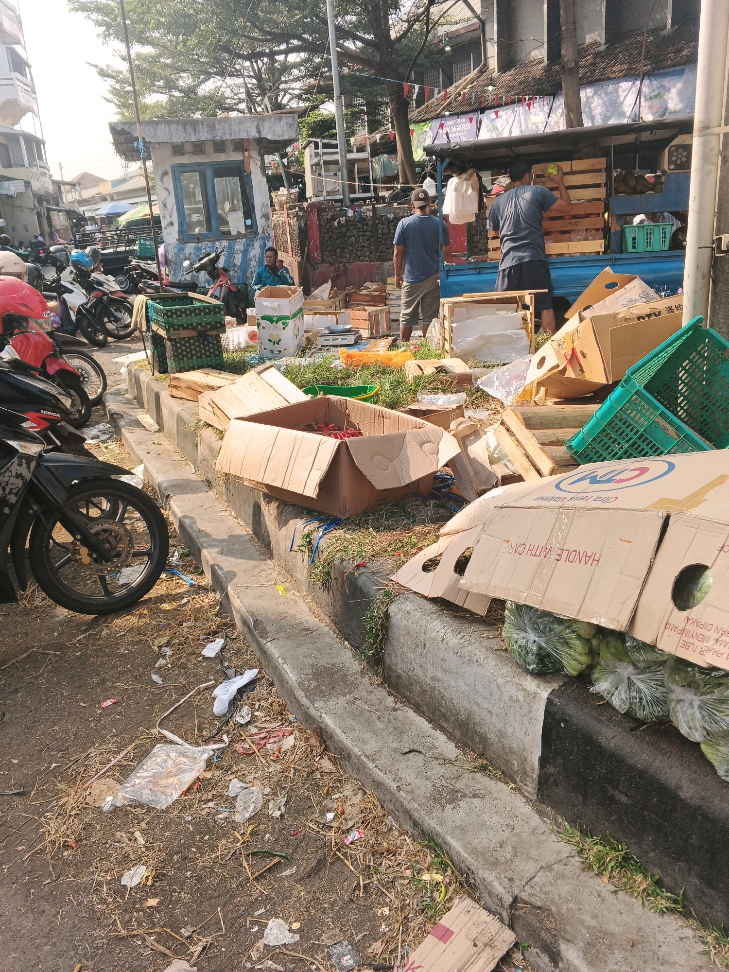 The image depicts a street scene in Ungaran, Indonesia, likely a market area due to the presence of goods and transaction activities. The foreground shows a sidewalk cluttered with debris, including dry grass, plastic bags, and paper. Several motorcycles are parked along the curb. A prominent element is a collection of cardboard boxes, some open and containing items, others stacked. Green plastic crates are also visible. In the middle ground, a small, weathered booth with blue trim stands by the roadside. Behind it, a blue truck is laden with wooden crates, and two men are interacting with the produce. One man, wearing a blue t-shirt and shorts, is holding a green item, possibly a fruit or vegetable, and appears to be placing it into a crate. Another man in a blue t-shirt and light-colored shorts stands facing away from the camera. The background reveals more buildings and trees, suggesting an urban or semi-urban environment. The overall impression is one of a busy, outdoor market with goods being handled and displayed.
