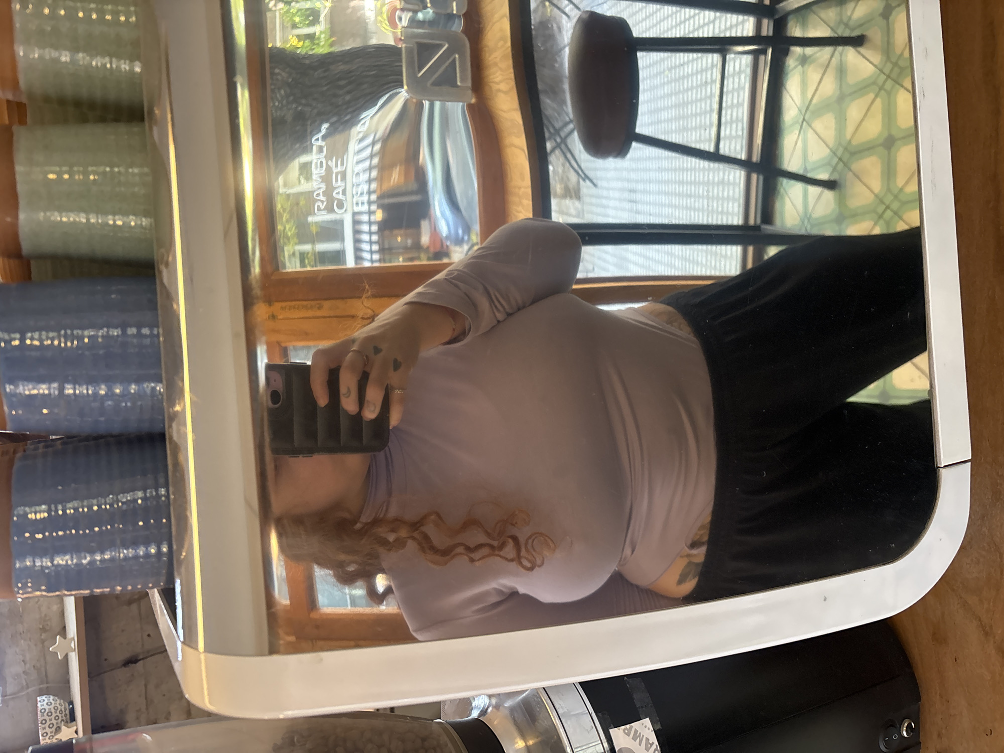 A person with curly brown hair is taking a mirror selfie with a smartphone. The mirror is rectangular with rounded corners and a white frame. In the reflection, the person is wearing a light purple turtleneck shirt and black pants. The reflection also shows a portion of "RAMBLA CAFE ESPIRITU," a chair, and a tiled floor. Outside the mirror's reflection, stacked cylindrical containers of varying colors are visible, along with a coffee grinder. The location is Buenos Aires, Argentina.