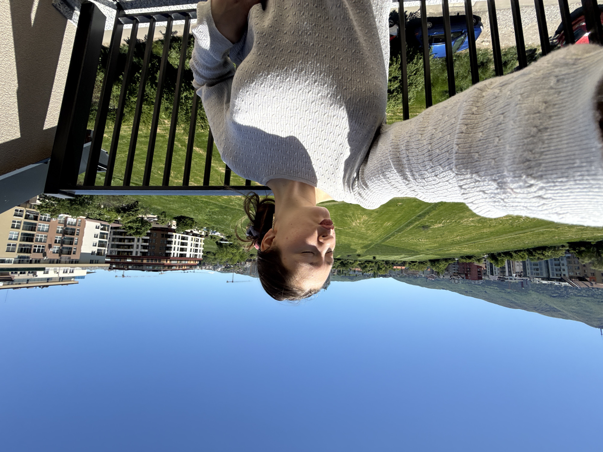 A woman is taking a selfie on a balcony in Sofia, Bulgaria. She is wearing a light-colored sweater and has her eyes closed. Behind her are buildings, a large grassy field, and a mountain range. A blue car and a red car are visible on the road. The sky is clear and blue. The balcony has black metal railings.