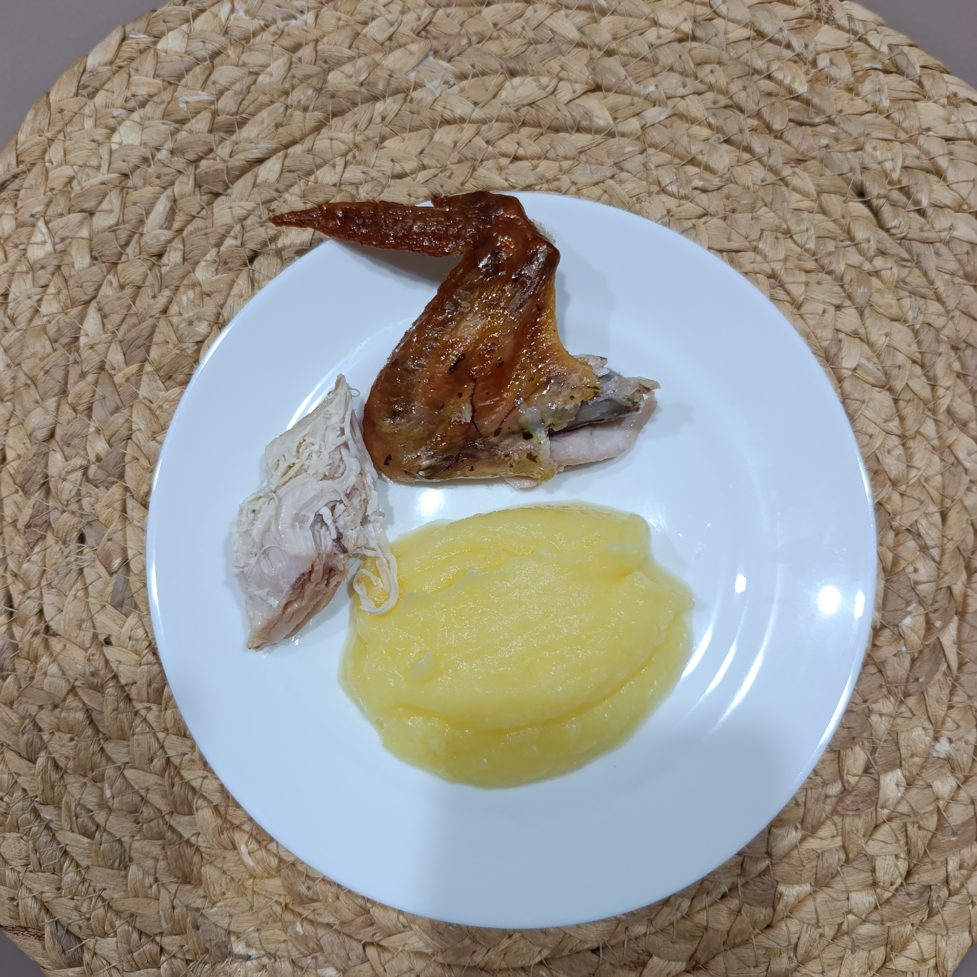 A white, circular ceramic plate is centrally positioned on a woven, light brown, natural fiber placemat. The plate holds a meal consisting of three components: an intact, roasted poultry piece, likely a wing or drumette, displaying a glossy, browned, crispy skin; a smaller, irregularly shaped portion of shredded white poultry meat adjacent to the roasted piece; and a roughly oval-shaped mound of smooth, yellow mashed potatoes or a similar purée. The background is composed entirely of the woven placemat, which exhibits a consistent, tight basket weave pattern. The scene depicts a prepared meal, possibly in a domestic setting, in the city of Марковский, Russia.