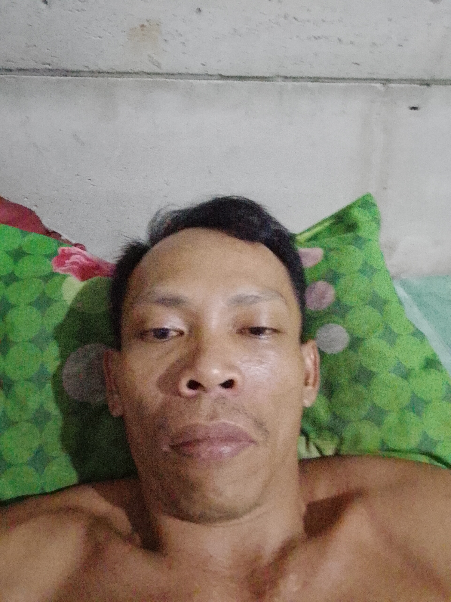 A man lies on a bed with green and pink patterned pillows. He appears to be taking a selfie. The wall behind the pillows is concrete. The image provides no information to verify the location in Bukit Ketok, Indonesia.