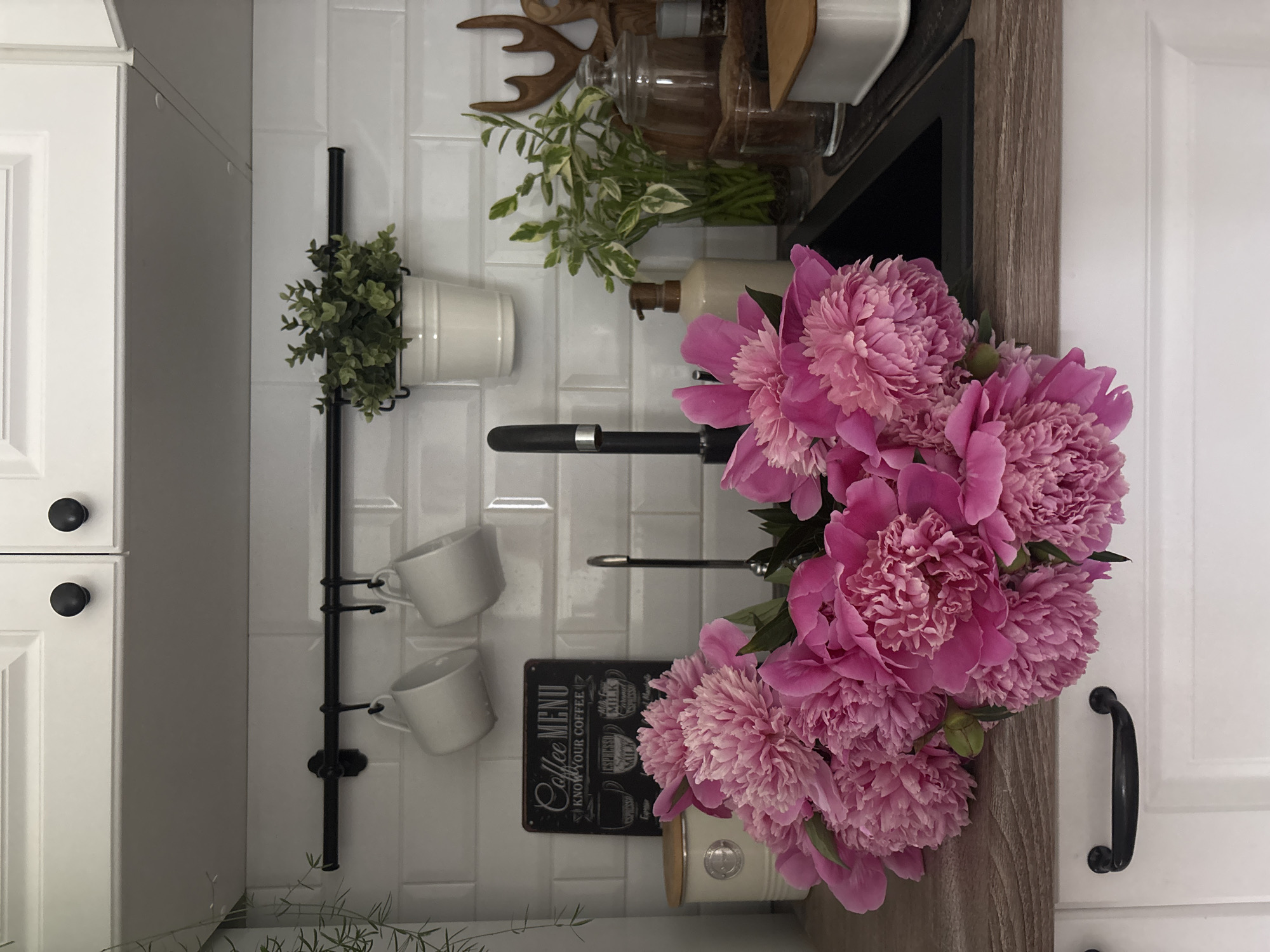 A close-up shot captures a bouquet of vibrant pink peonies resting on a wooden countertop in a modern kitchen. The flowers, in full bloom, cascade over the edge of the counter, their ruffled petals creating a lush, textural display. Behind the peonies, a black kitchen sink is visible, with a sleek black faucet rising from its center. To the left of the sink, a small, vintage-style sign with "Coffee MENU" written on it hangs on a white subway-tiled backsplash. Above the sign, two white ceramic mugs are suspended from a black metal rack, alongside a small potted green plant. The kitchen features white cabinetry with black hardware. The scene suggests a domestic setting, likely a home kitchen, in the city of Chaikovsky, Russia.