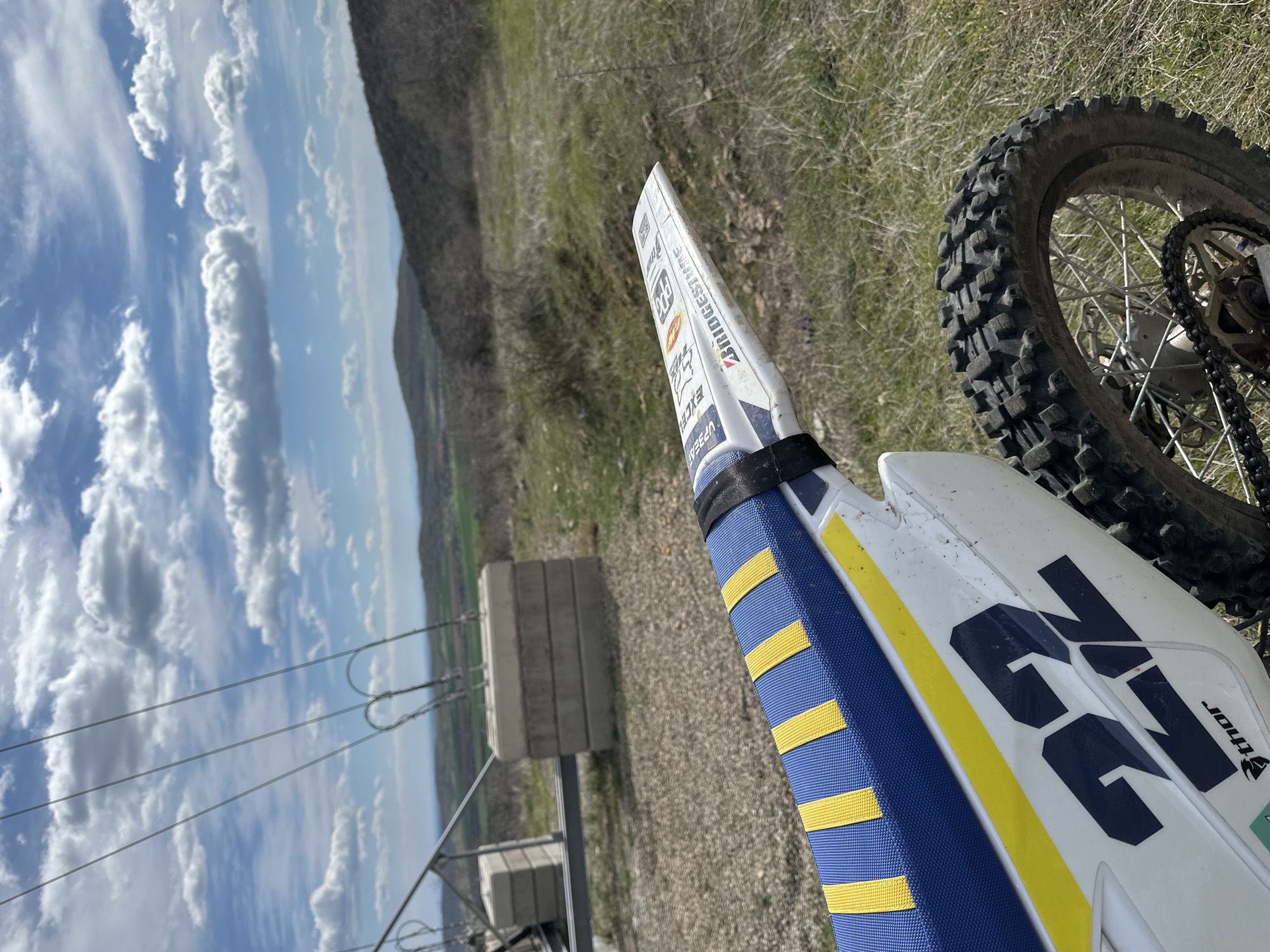 The image shows a dirt bike in a rural area, potentially near electrical infrastructure. The bike's front fender has stickers from Bridgestone and other brands. The seat is blue with yellow stripes, and the side panel has the number 22 on it. The rear tire is visible, with a knobby tread. In the background, there's a grassy hill, some agricultural land, and a partly cloudy sky. A utility pole with power lines is also in the background. The location is in the city of Unknown, Unknown.