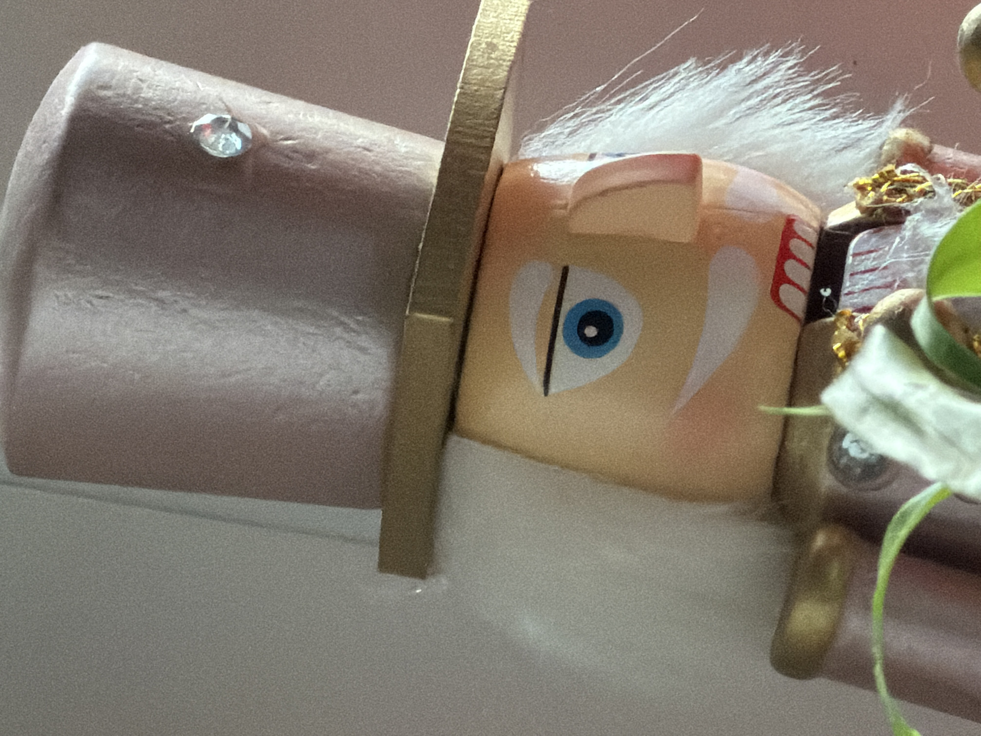 Close-up of a decorative wooden nutcracker, partially in focus. The nutcracker has a light brown face with a single, large blue eye. It wears a tall, cylindrical hat with a rhinestone, and a gold band. White hair is visible on top. The nutcracker appears to be part of a larger object and is tilted horizontally. The background is a neutral color. There are no people visible in the image. There is no information available to determine its location in Esch-sur-Alzette, Luxembourg.