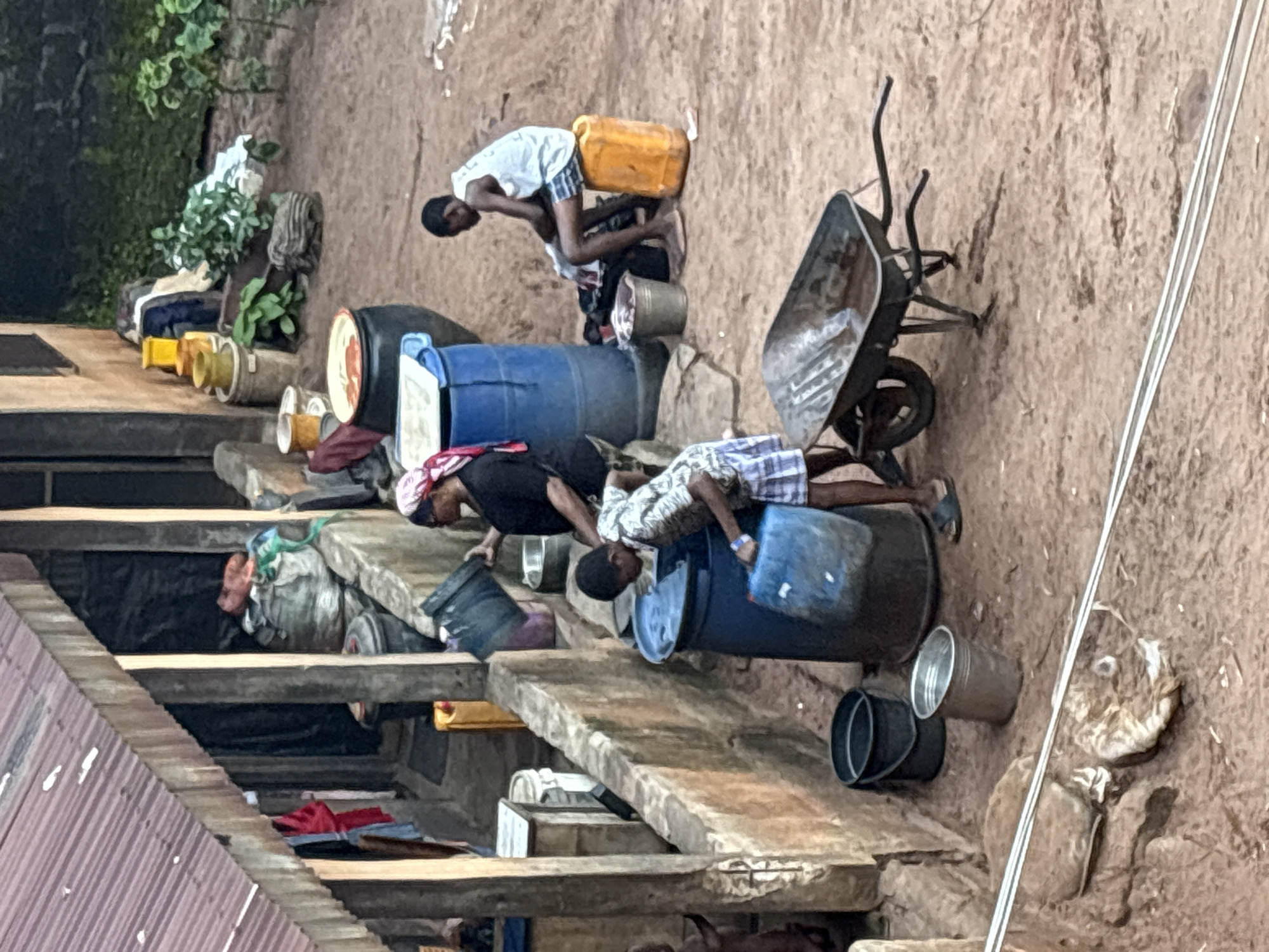 In Onitsha, Nigeria, a scene of daily life unfolds outside a dwelling. A corrugated iron roof extends over a covered area where various items are stored, including large sacks, buckets, and what appear to be cooking pots. A woman wearing a headscarf and black top is seen tending to a large blue barrel, holding a blue container. Nearby, a young boy in a patterned shirt and shorts is also interacting with the blue barrel, carrying a blue plastic container. Further to the right, another young boy sits on an orange plastic container, wearing a white tank top and shorts, with some clothing and bags around him. In the foreground, a wheelbarrow filled with water is positioned on the dusty, uneven ground. Several dark buckets and a metal bucket with water are also present on the ground. The environment suggests a warm climate with dry, reddish-brown soil.