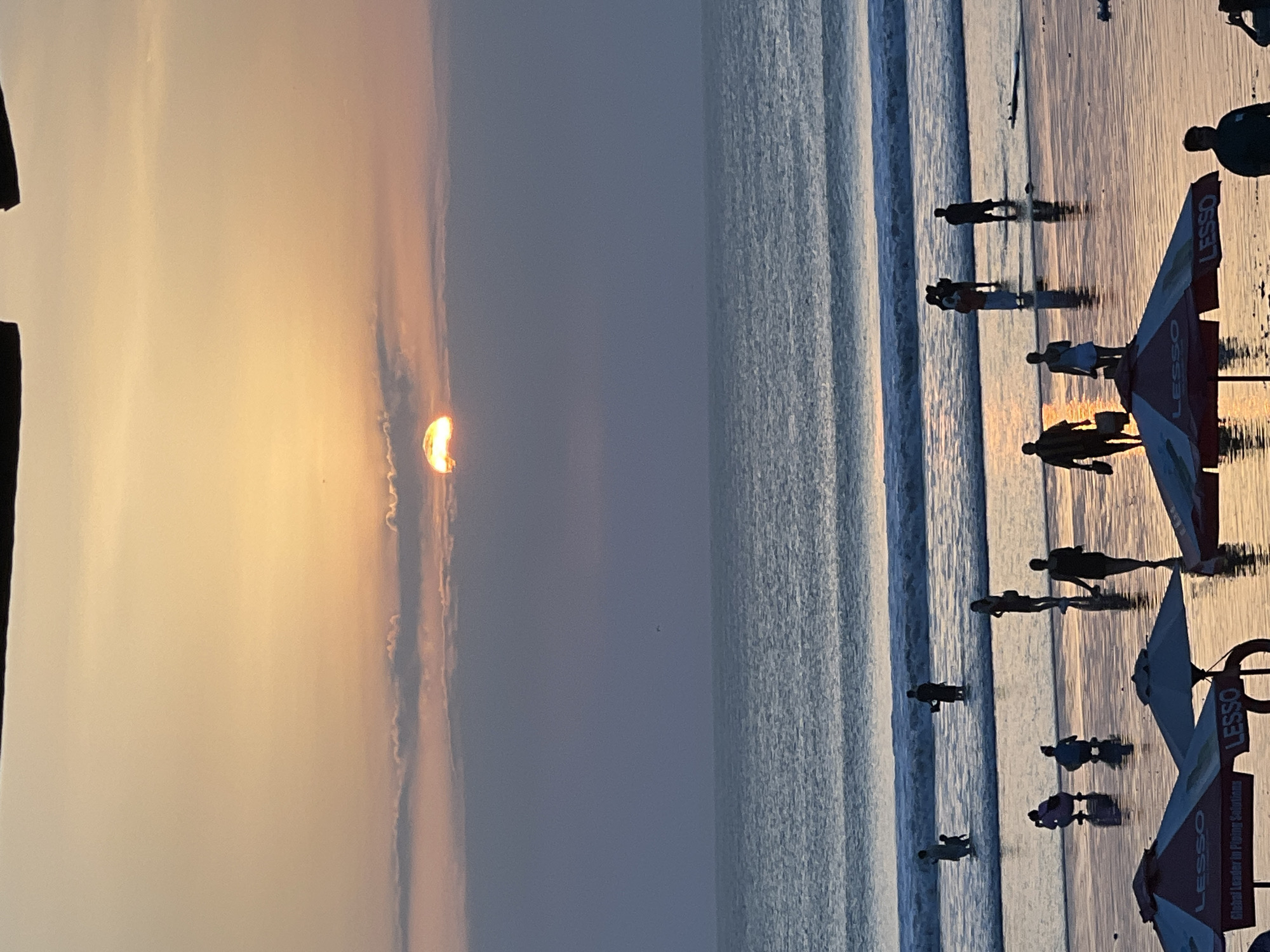 A wide shot captures a sunset scene at a beach in Kuta, Indonesia. The sun, partially obscured by clouds, casts a warm orange glow across the sky and reflects on the wet sand and ocean. Several people are visible on the beach, some wading in the shallow water, others standing or walking on the sand. Beach umbrellas are set up in the foreground, with some advertising the brand "LESSO." The ocean has gentle waves rolling towards the shore. The overall atmosphere appears serene and peaceful as the day transitions to evening.