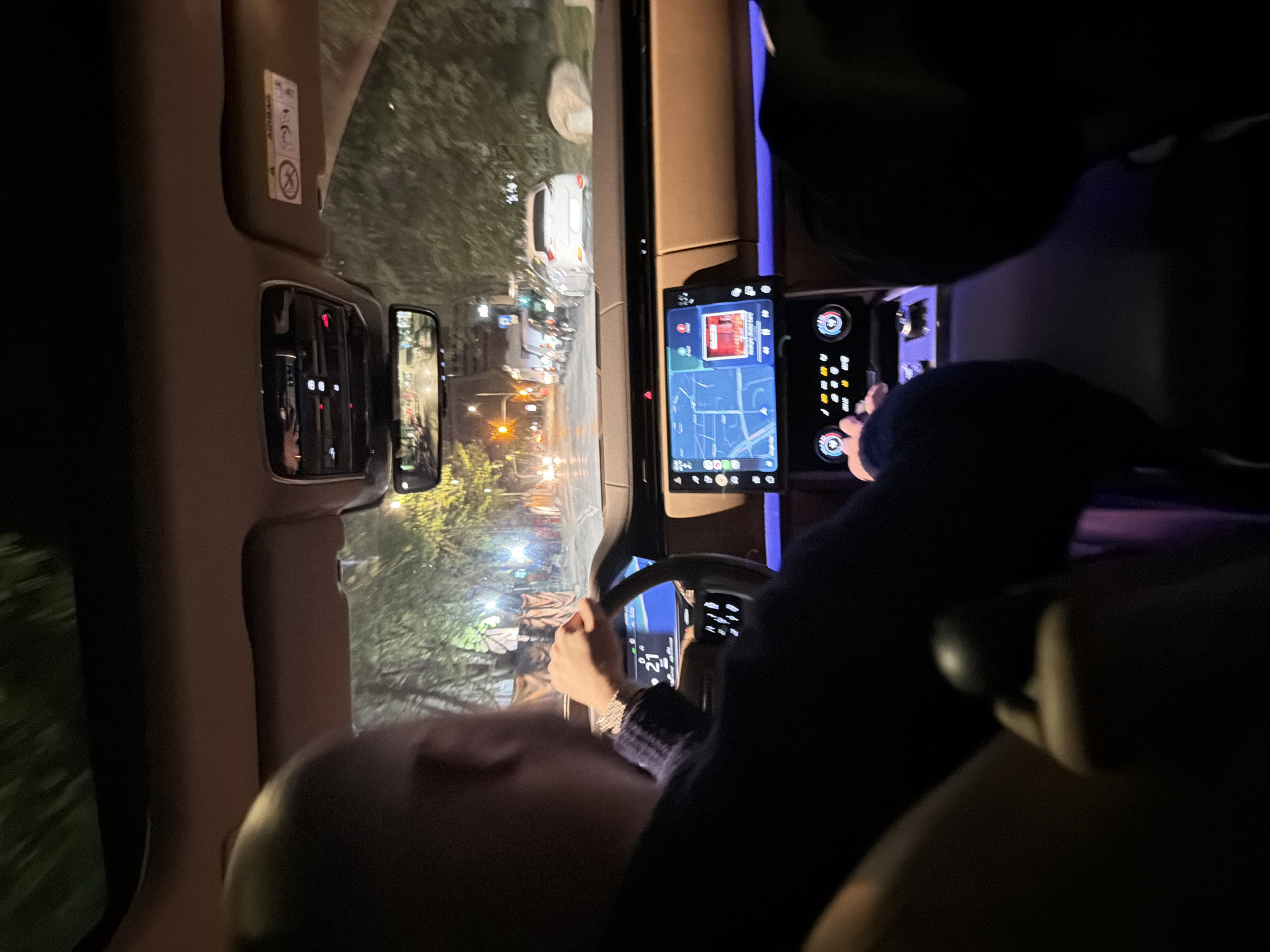 The image shows the interior of a car at night, with the view looking out the windshield onto a city street. Car headlights and streetlights illuminate the scene. A car is visible in front, with other vehicles further down the road. Inside the car, the driver's hands are visible on the steering wheel, and the center console screen shows a map. There is a passenger in the car. The location is Unknown, Unknown.