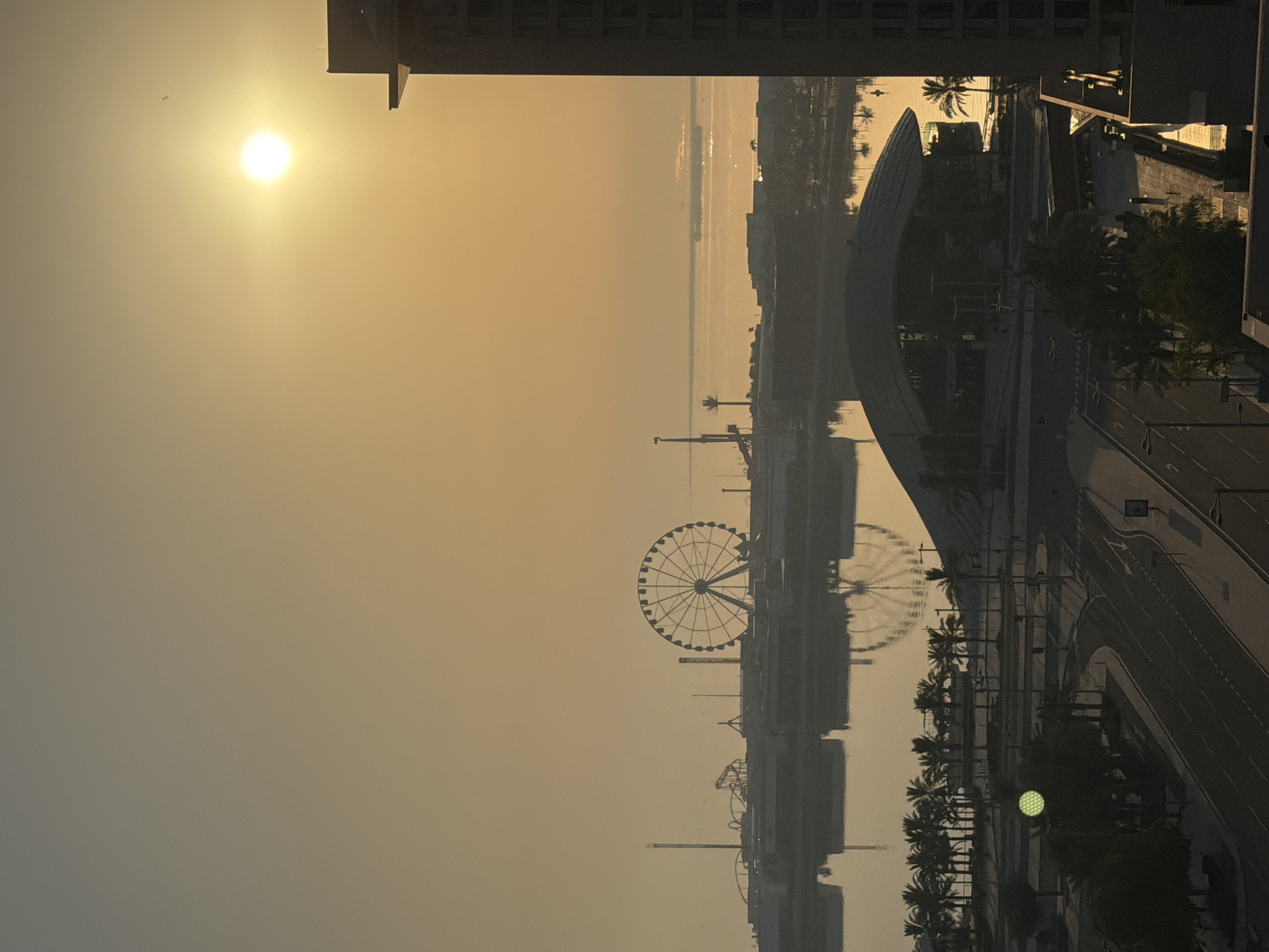 This is an aerial, high-angle, eye-level, medium shot of a coastal city at sunrise. The sun is a bright orb in the hazy sky, casting a warm, golden glow over the scene. In the foreground, a dark, modern building dominates the right side of the frame, its geometric structure silhouetted against the sunrise. Below it, a street curves through a city landscape dotted with palm trees and other vegetation. A large, futuristic-looking building with a curved roof is visible in the middle ground, its dark form contrasting with the bright water behind it.
Beyond the city, a body of water reflects the sky's golden light, with a large ship visible in the distance. On the left side of the frame, a Ferris wheel and other amusement park rides stand as dark silhouettes against the horizon, hinting at a recreational area. The overall impression is one of a serene, awakening city bathed in the soft light of dawn.