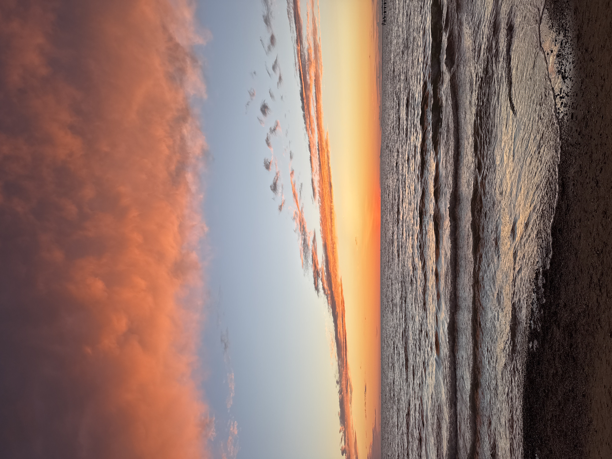 This is a vibrant landscape photograph depicting a sunset or sunrise over a coastal area in Anapa, Russia. The image captures a broad view of the sky, sea, and shoreline, with the horizon line prominent across the frame.
The sky, occupying the upper portion of the image, displays a dramatic array of colors. On the left, a large, expansive cloud mass glows in intense shades of orange and reddish-pink, illuminated by the low sun. Towards the center and right, the sky transitions from a soft, clear blue directly above the horizon to warm hues of golden yellow and bright orange where the sun is setting or rising. Scattered, elongated clouds, also touched by the sun's warm light, are visible across the middle sky.
Below the horizon, the calm sea stretches into the distance, reflecting the fading light. The water's surface shows gentle ripples and waves, catching the light and creating a textured pattern of highlights and shadows. The foreground, particularly on the right side, reveals a dark, wet sandy beach or shoreline, textured with the receding water lines and possibly some foam.
In the far distance, near the horizon line on the right, a faint structure resembling a pier or jetty extends into the water, silhouetted against the bright sky. No people or specific activities are visible, emphasizing the serene and expansive natural scene. The image is devoid of any visible text. The overall atmosphere is one of tranquility and natural beauty, characteristic of a coastal twilight.