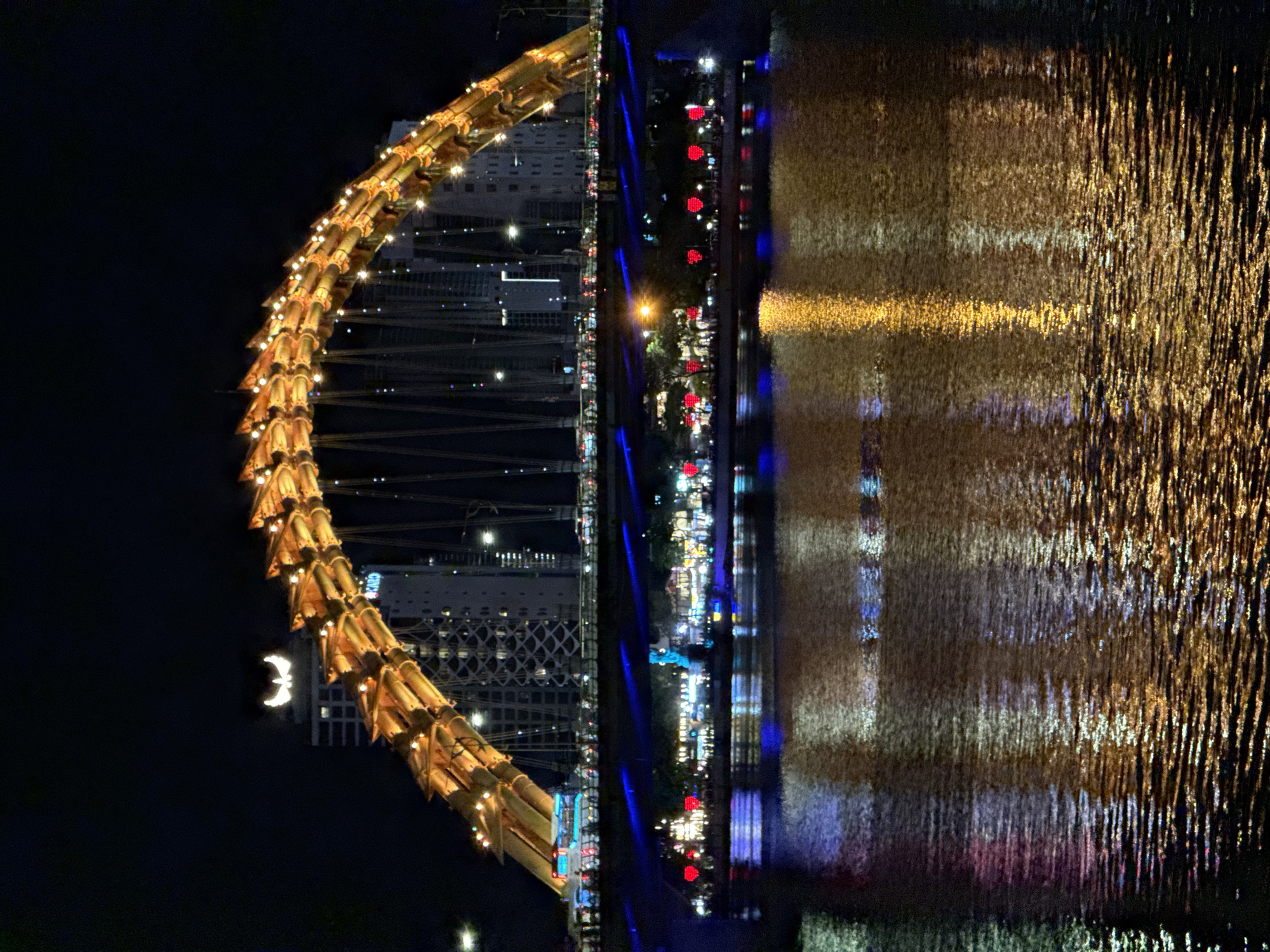This nighttime image displays the Dragon Bridge over the Han River in Đà Nẵng, Vietnam. The bridge's main structure, an arch designed to resemble a dragon, is centrally positioned and illuminated with a golden-orange light emanating from numerous individual fixtures along its length. Directly behind the dragon's head portion of the bridge, on the facade of a tall building, a prominent white, winged emblem resembling a crescent moon is visible. Below the main arch, the bridge's lower traffic deck is illuminated with blue and white lights, showing multiple vehicles, including cars and at least one bus, in transit. The riverbanks beneath the bridge feature various illuminated structures and a series of red decorative lights, some appearing heart-shaped. The Han River occupies the foreground, its rippled surface reflecting a dense array of golden, blue, and reddish lights from the bridge and the adjacent urban environment, creating elongated, shimmering patterns across the water.