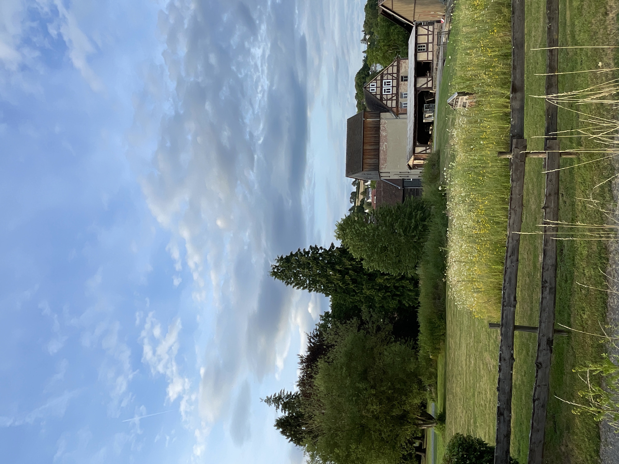 The image depicts a pastoral scene in Neukirchen, Germany. A rustic wooden fence runs diagonally across the right side of the frame, separating a lush green lawn from a field dotted with wildflowers. In the background, traditional German Fachwerk houses with timbered facades and white-paned windows are nestled amongst rolling green hills and mature trees. The sky above is a dynamic canvas of blue, streaked with wispy white clouds and darker, more dramatic grey formations, suggesting a partly cloudy sky. A faint airplane contrail is visible in the distance. The overall impression is one of a peaceful, rural setting, likely in the late afternoon or early evening, given the soft, diffused light. No people or specific activities are evident, but the scene evokes a sense of tranquility and the charm of the German countryside. The architecture of the houses and the rural landscape strongly indicate a European location, consistent with the provided location context.