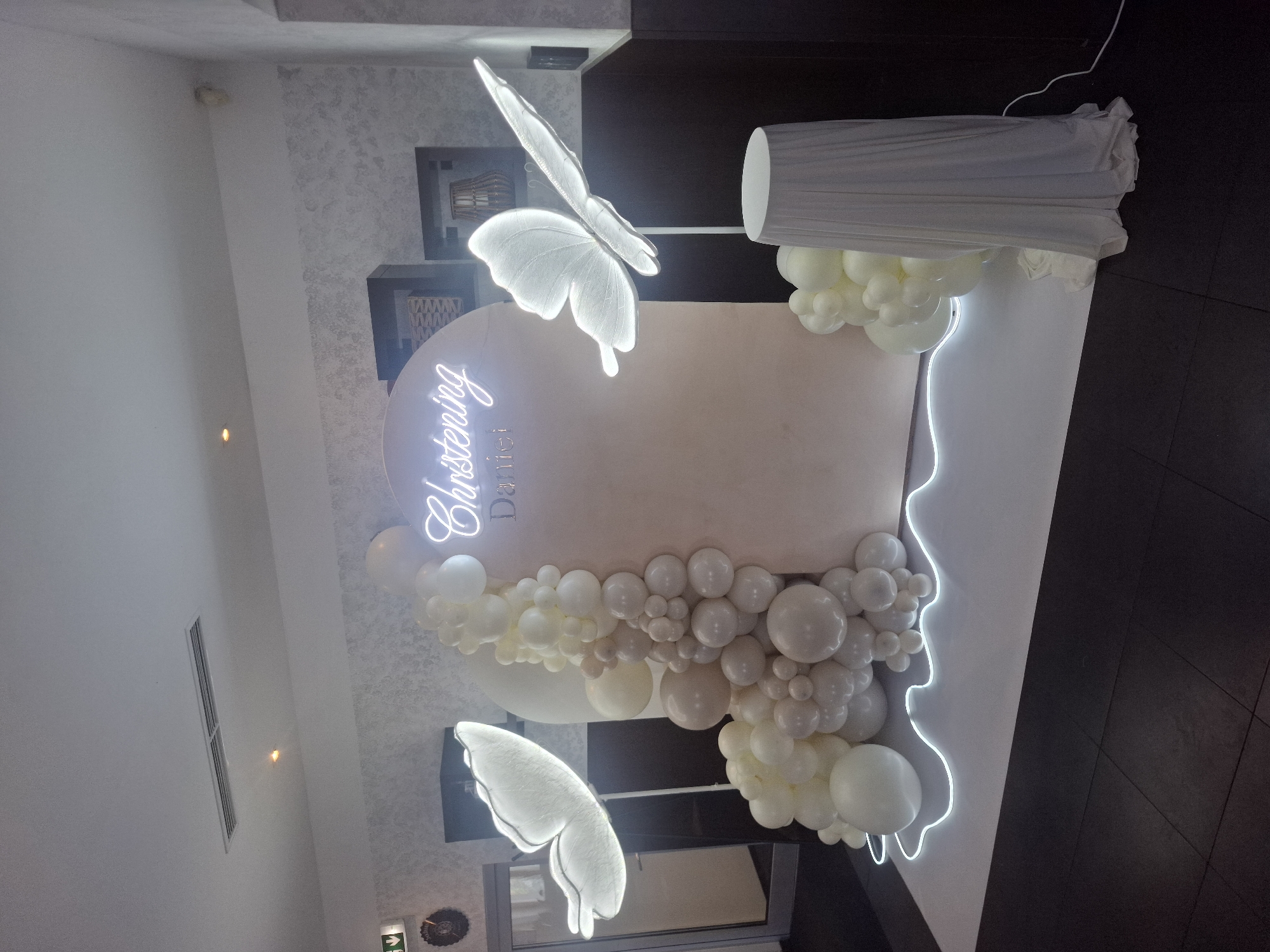 This is a full-shot, eye-level, indoor photograph of a christening setup. The scene features a circular backdrop with a neon sign that reads "Christening Daniel" in a cursive font. To the left and right of the backdrop are large, illuminated white butterfly decorations mounted on stands. A balloon garland in shades of white and cream is arranged around the base of the backdrop, extending to the left. Another cluster of balloons is placed to the right. A white draped table or pedestal is positioned in front of the right butterfly. The floor is tiled in a dark color, and the overall backdrop area is illuminated by white LED strip lights that outline the platform. The walls in the background have a textured wallpaper and dark paneling. There are no people visible in the image. This setup is likely for a celebration event in a venue, such as a restaurant or event hall.