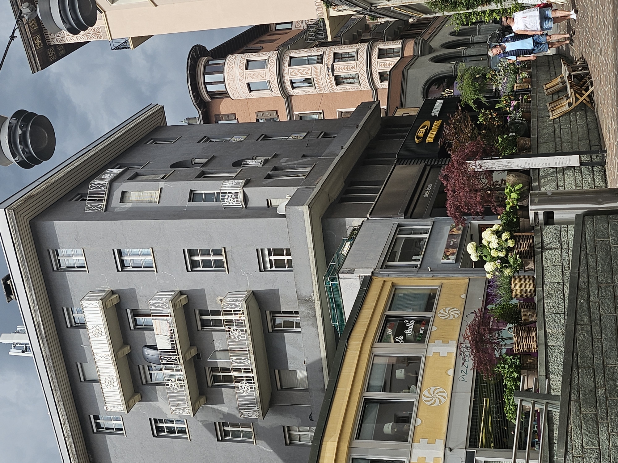 The image depicts a street scene in St. Moritz, Switzerland, featuring a blend of architectural styles. On the left, a grey, multi-story building with numerous windows and white-railed balconies dominates the foreground. Below this building, a yellow facade with large windows displays signs for "Pizza La Stalla Ristorante." In front of the yellow building, a row of potted plants, including a Japanese maple, hydrangeas, and lavender, are arranged on a stone wall.
To the right of the grey building, a distinctive round, pinkish-orange building with ornate white trim and arched windows stands prominently. Further to the right, another building with arches and dark grey facades is visible, housing shops with signage. Two individuals, a man and a woman, are walking on the cobblestone sidewalk, with the man wearing a blue vest and shorts, and the woman in a white top and shorts. Two wooden chairs are positioned on the sidewalk in front of the buildings. The sky is overcast with dramatic clouds. Overhead, two large, industrial-looking light fixtures are suspended.