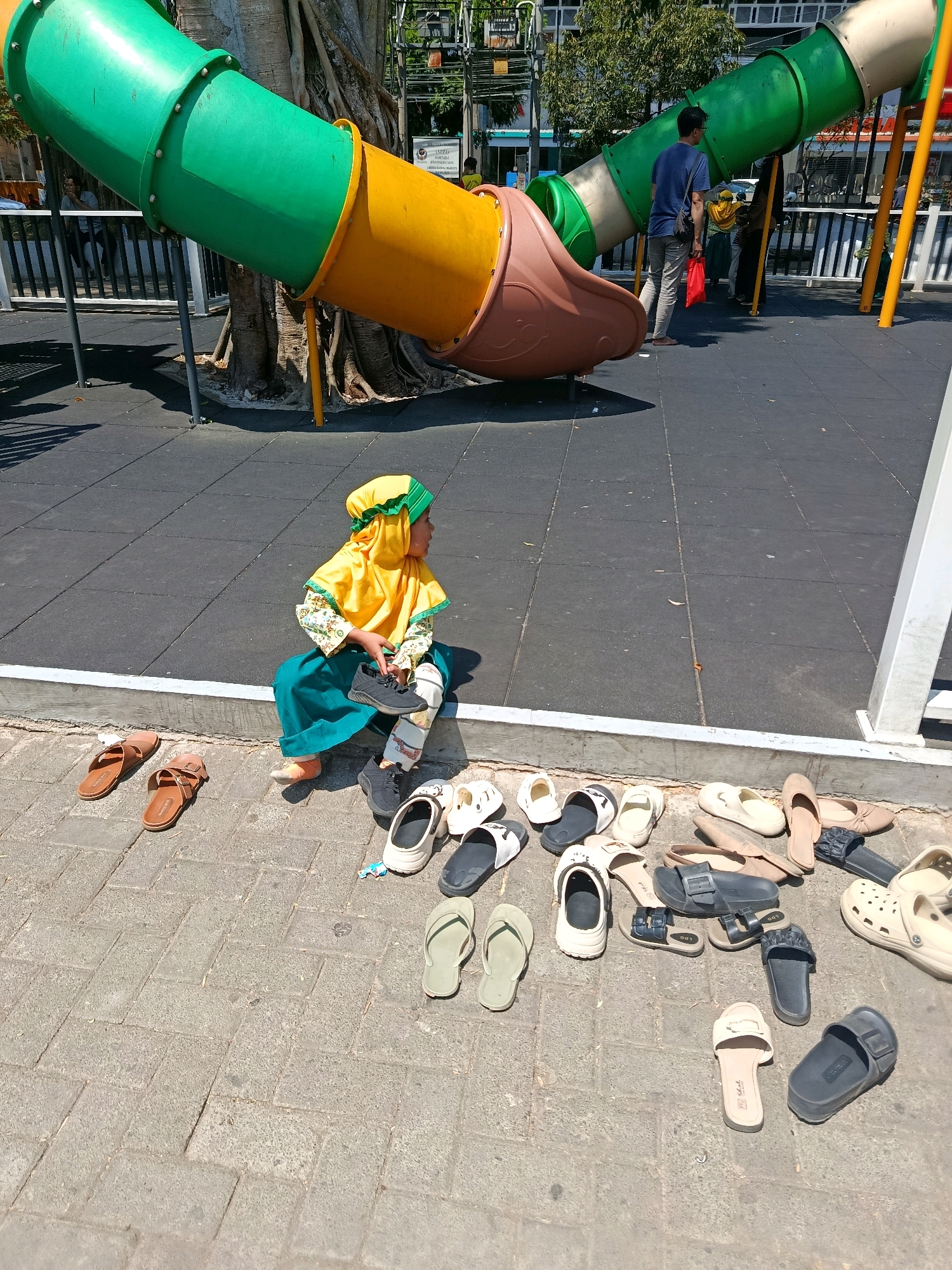 In a Malang, Indonesia public park, a young child dressed in a yellow headscarf and green skirt sits on a paved area next to a children's playground slide structure. The child holds a dark-colored shoe in their hands. Numerous pairs of footwear, including sandals and slippers, are scattered on the ground around the child. In the background, adults are visible near the playground equipment, and trees and buildings suggest an urban park setting. The ground is a mix of dark rubber matting under the playground and light grey paving stones.