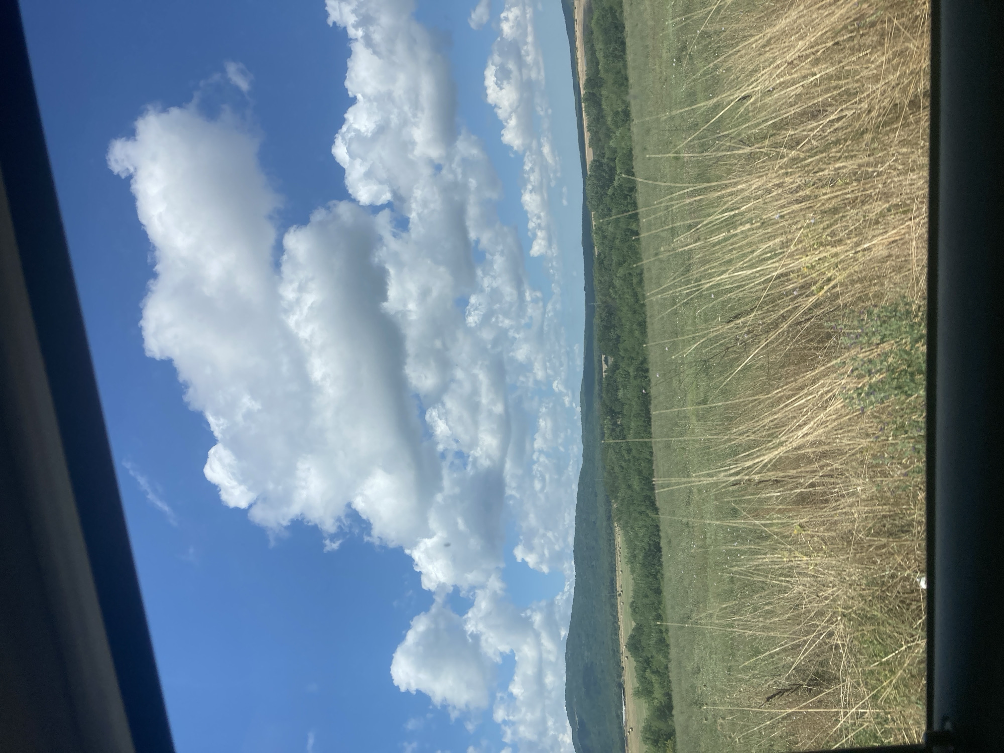 The image captures a pastoral landscape under a bright blue sky dotted with puffy white clouds. In the foreground, tall, dry grasses dominate the scene, their stalks reaching upwards. Beyond the grasses, a band of green trees stretches across the middle ground, with rolling, forested hills forming the background. The sky is expansive, with large cumulus clouds casting shadows on the landscape below. There are no people or animals visible in the frame. The perspective suggests the photo was taken from within a vehicle, looking out through a window. The image depicts a rural environment, likely outside the town of Ravadinovo, Bulgaria, given the mention of this location.