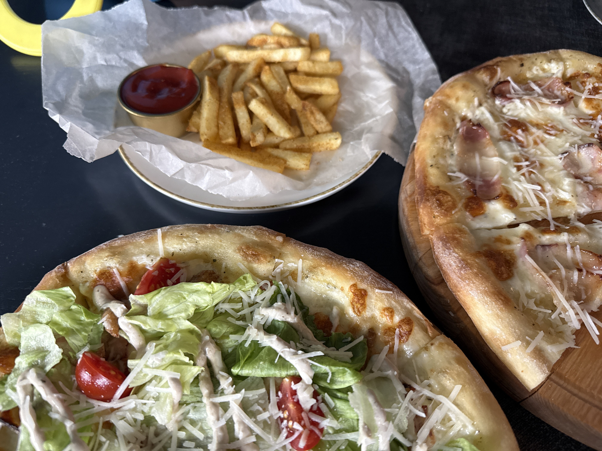 The image displays a close-up, overhead view of a food spread on a dark, textured surface, likely a table. In the foreground, occupying the bottom right quadrant, is a slice of pizza on a wooden board. This pizza appears to have a white cheese base with visible pieces of what could be ham or bacon, and is sprinkled with grated cheese. To the left and slightly behind this slice is another pizza, larger and circular, with a crust showing browning. Its toppings include shredded lettuce, halved cherry tomatoes, and a creamy dressing, resembling a Caesar salad pizza.
To the right of the pizzas, in the upper portion of the image, is a white paper liner within a shallow bowl or plate. This liner holds a serving of golden-brown french fries, some of which are dusted with seasoning. A small, round metal ramekin filled with bright red ketchup sits atop the fries, partially obscuring them. The overall impression is a casual meal setting.
The location provided, Балаклавский округ, Ukraine, does not have specific visual cues within the image to confirm or deny. The scene itself is domestic or restaurant-oriented, focusing solely on the prepared food items. There are no people or animals visible. The lighting suggests an indoor environment, possibly with natural light coming from the side, casting soft shadows. The background is largely out of focus, dominated by the dark surface.