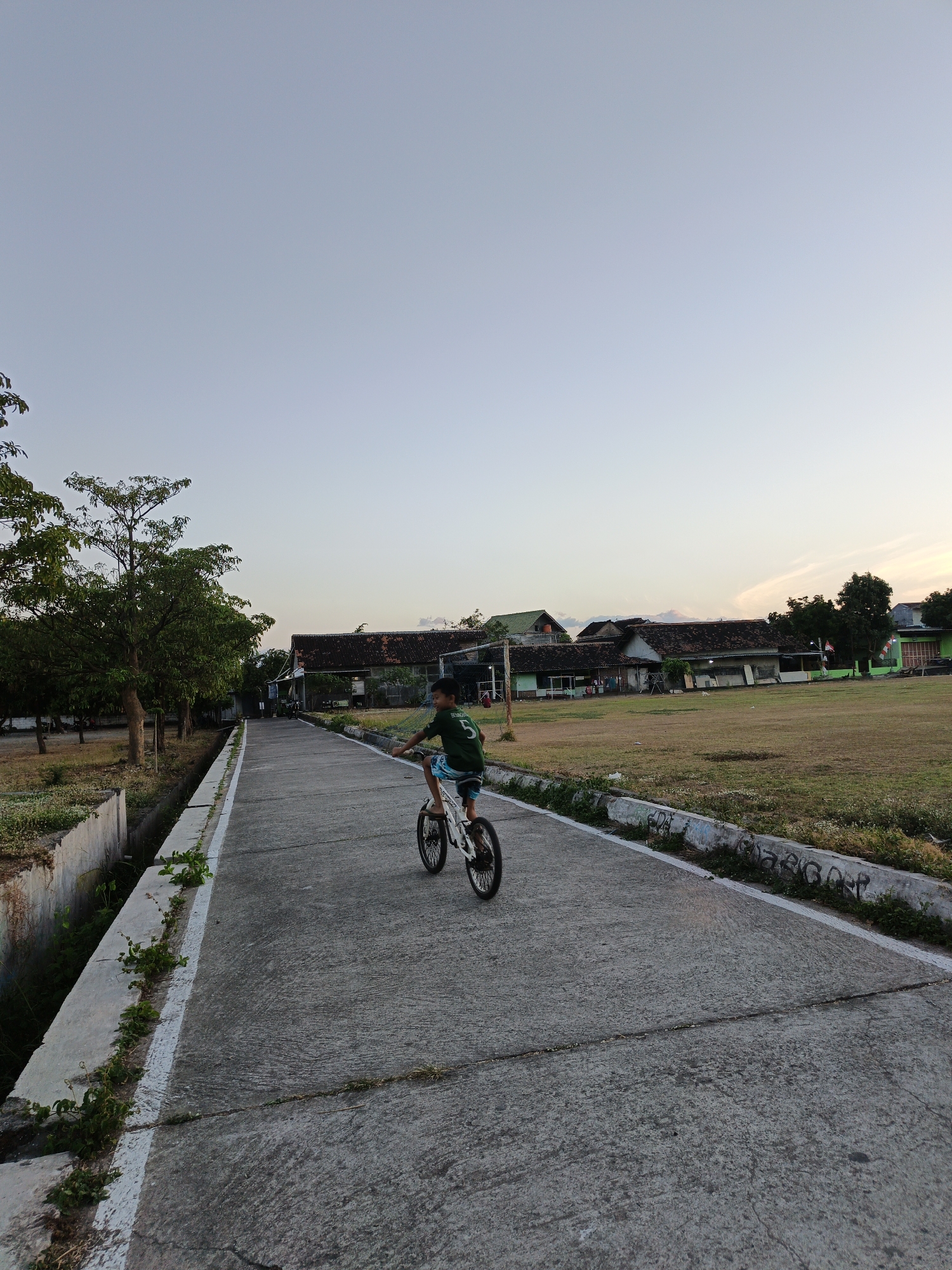 A young boy rides a bicycle down a concrete path in a rural setting in Klaten, Indonesia. The boy is wearing a green shirt with a number 5 on the back and blue shorts. He is positioned in the middle of the path, facing away from the camera. To the left of the path is a concrete drainage ditch with some overgrown vegetation. To the right of the path is a grassy field. In the background, there are several buildings with tiled roofs, likely houses or school buildings. Trees are visible on the left side of the path. The sky is clear and light blue, suggesting it is either early morning or late afternoon. The overall scene depicts a peaceful, everyday moment in a suburban or rural Indonesian environment.