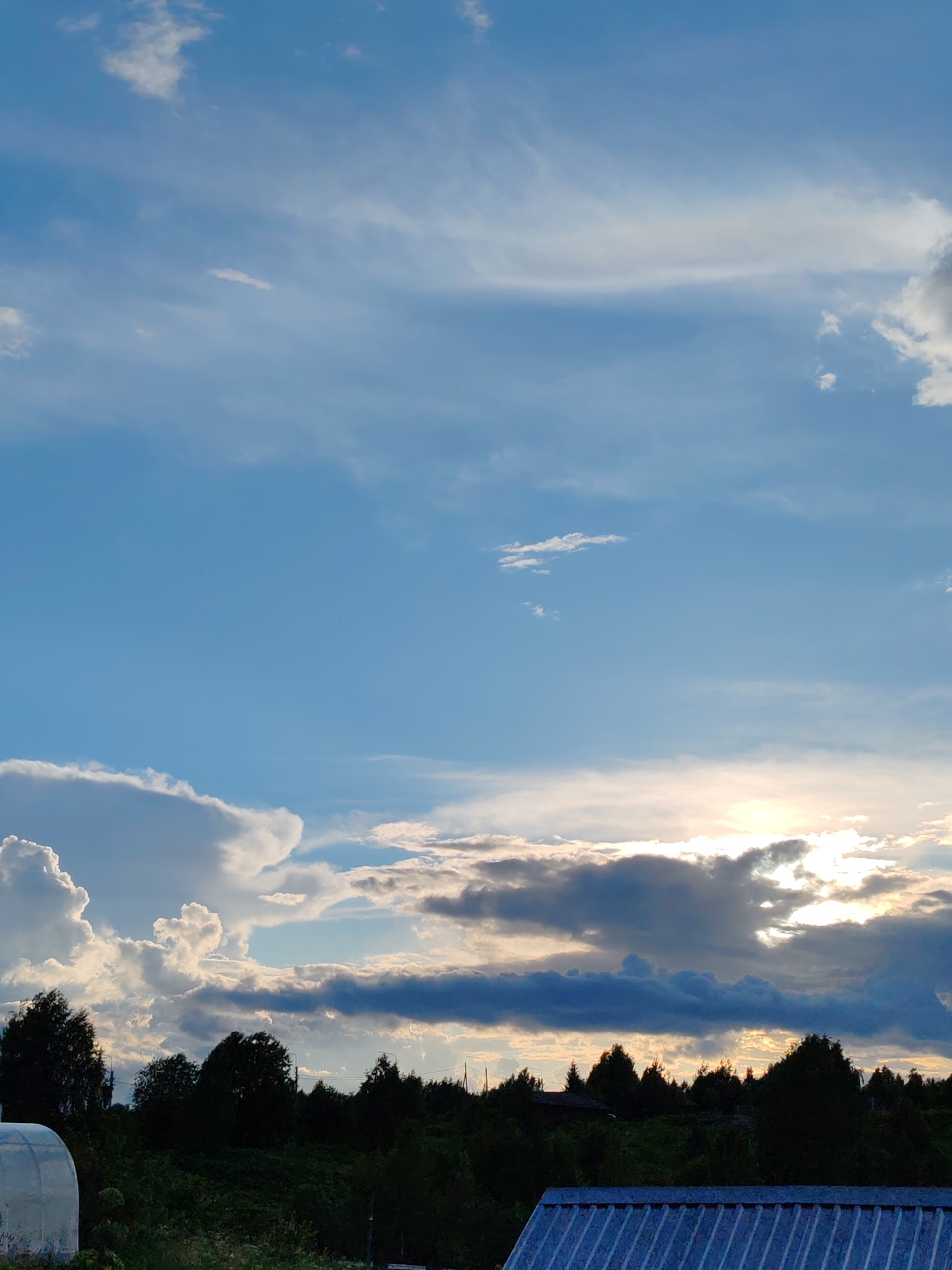 This vertical, medium-shot photo captures a picturesque rural landscape under a dramatic sky in Chukhlem, Russia. The scene is dominated by a vast expanse of sky, transitioning from a clear, bright blue at the top to a more turbulent display of clouds closer to the horizon. Wispy, white clouds drift across the upper blue, while lower down, heavier, darker clouds gather, some tinged with the golden light of the setting sun. The sun itself is partially obscured by these clouds, creating a bright, radiant glow that pierces through the gloom.
Below the sky, a dense line of dark green trees forms a silhouette against the horizon, punctuated by the occasional glimpse of buildings or structures peeking through. In the foreground, the edge of a corrugated metal roof, possibly from a shed or outbuilding, is visible at the bottom right. To the left, a portion of a white, domed structure, likely a greenhouse or a similar agricultural building, is also present. The overall impression is one of natural beauty and tranquility, with the dynamic sky suggesting a change in weather or the approach of evening. The time of day is late afternoon or early evening, indicated by the low angle of the sun and the quality of light. There is no visible text in the image.