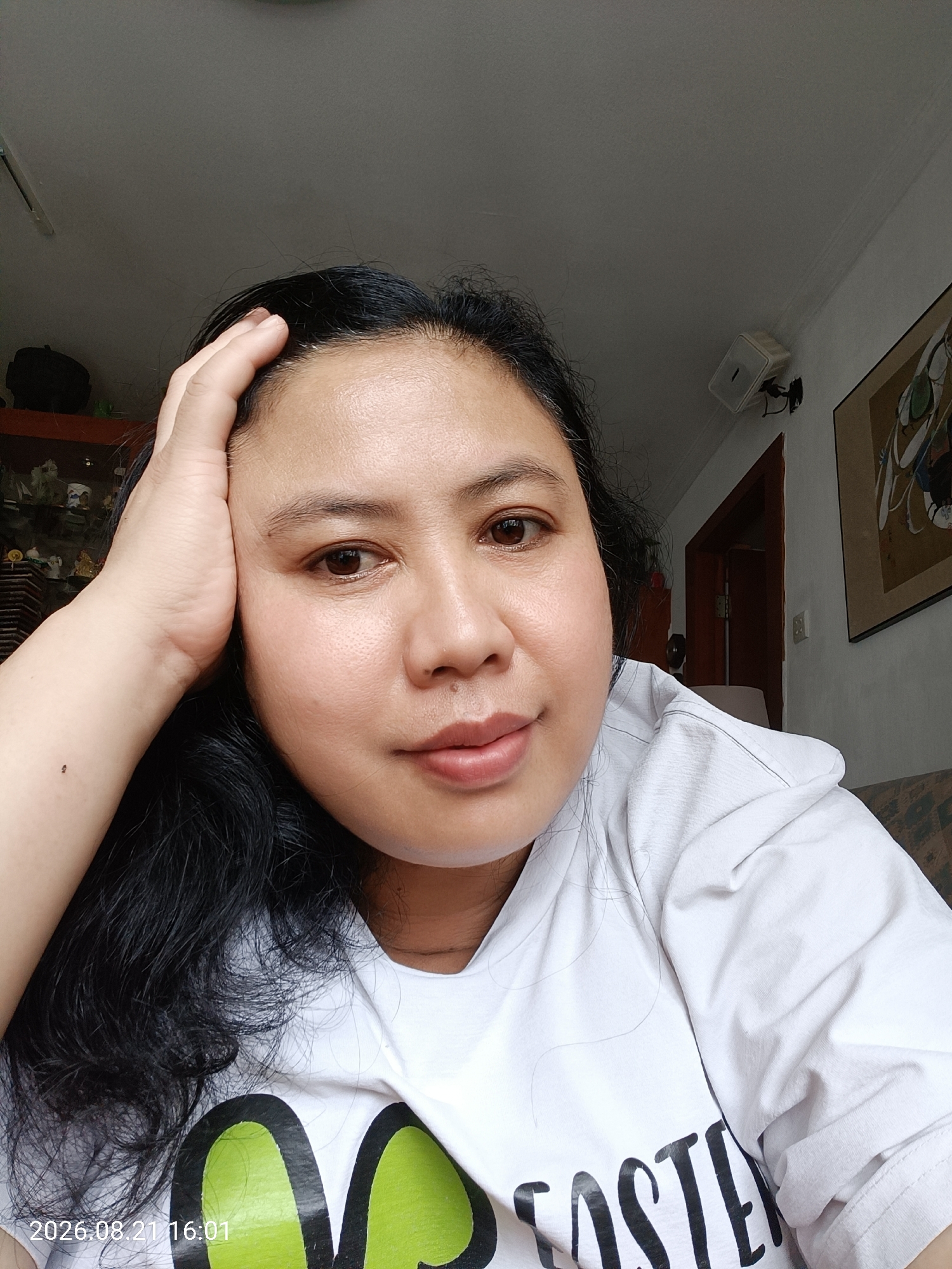 A medium shot captures a woman indoors, resting her forehead with her right hand. She has dark hair and is wearing a white t-shirt. The background shows a ceiling, a wall with a framed picture and a white speaker, and a doorway. A timestamp at the bottom left corner reads "2026.08.21 16:01". The location is not specified in the image.
